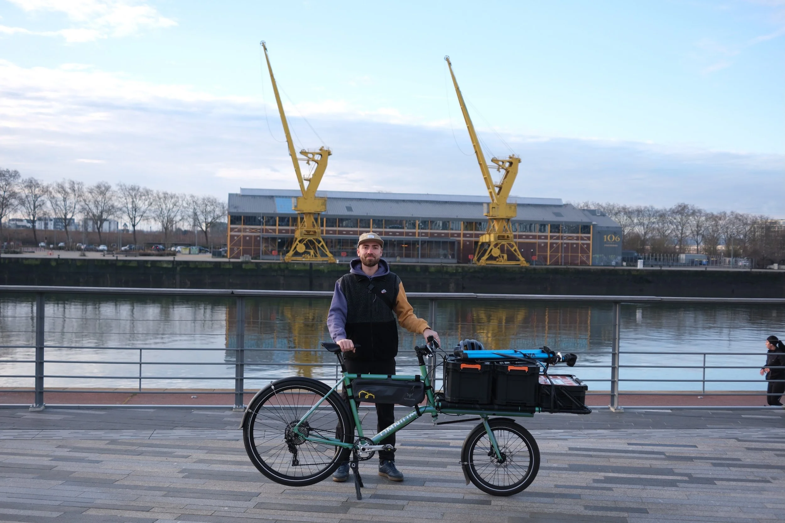 Homme avec un vélo cargo électrique garé devant une rivière, avec deux grues jaunes et un bâtiment en arrière-plan.