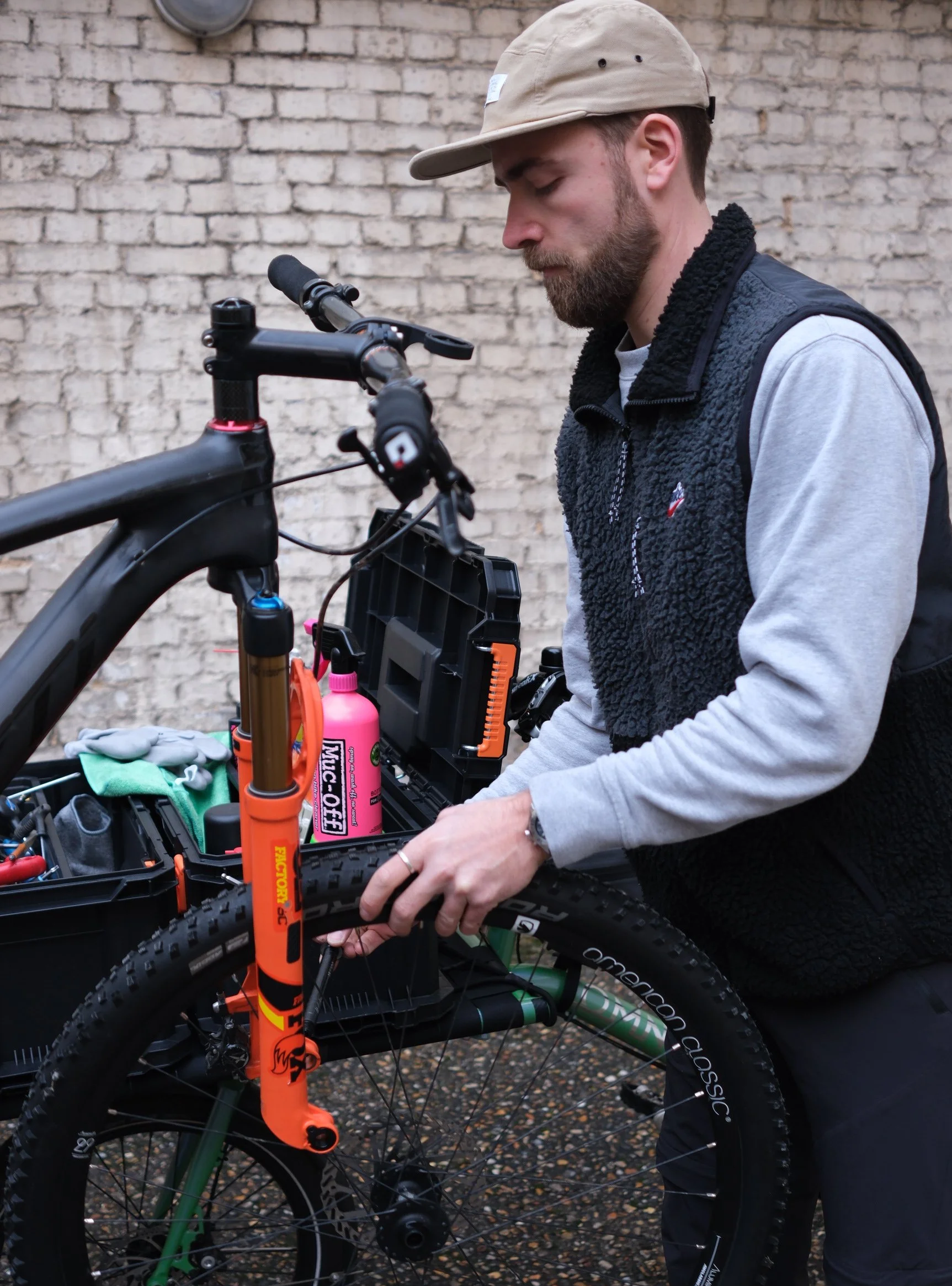 Un homme prépare un vélo électrique noir et vert avec un kit d'outils et un spray pour entretien devant un mur de briques.