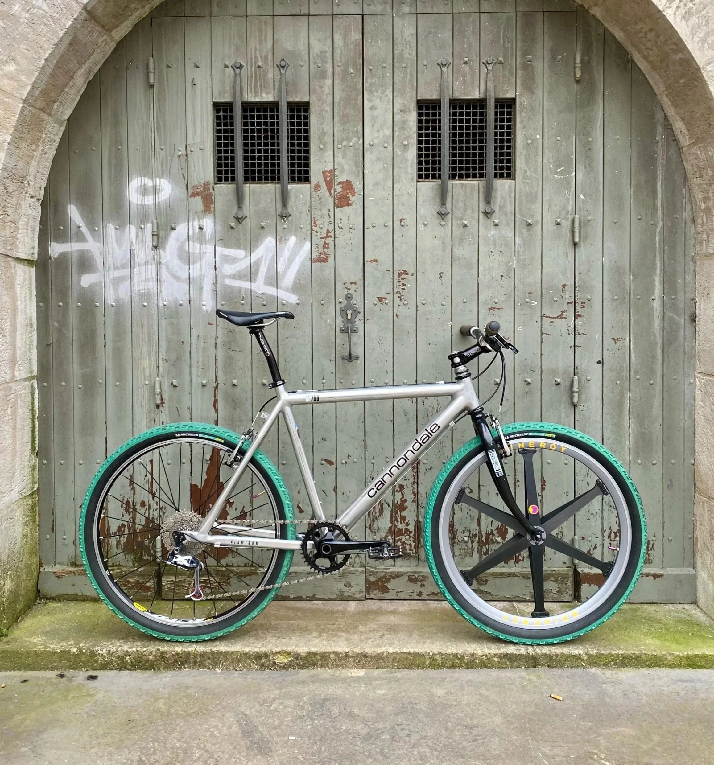 Vélo de montagne Cannondale avec pneus verts, en position verticale devant une porte en bois grise avec graffiti blanc et petites fenêtres.