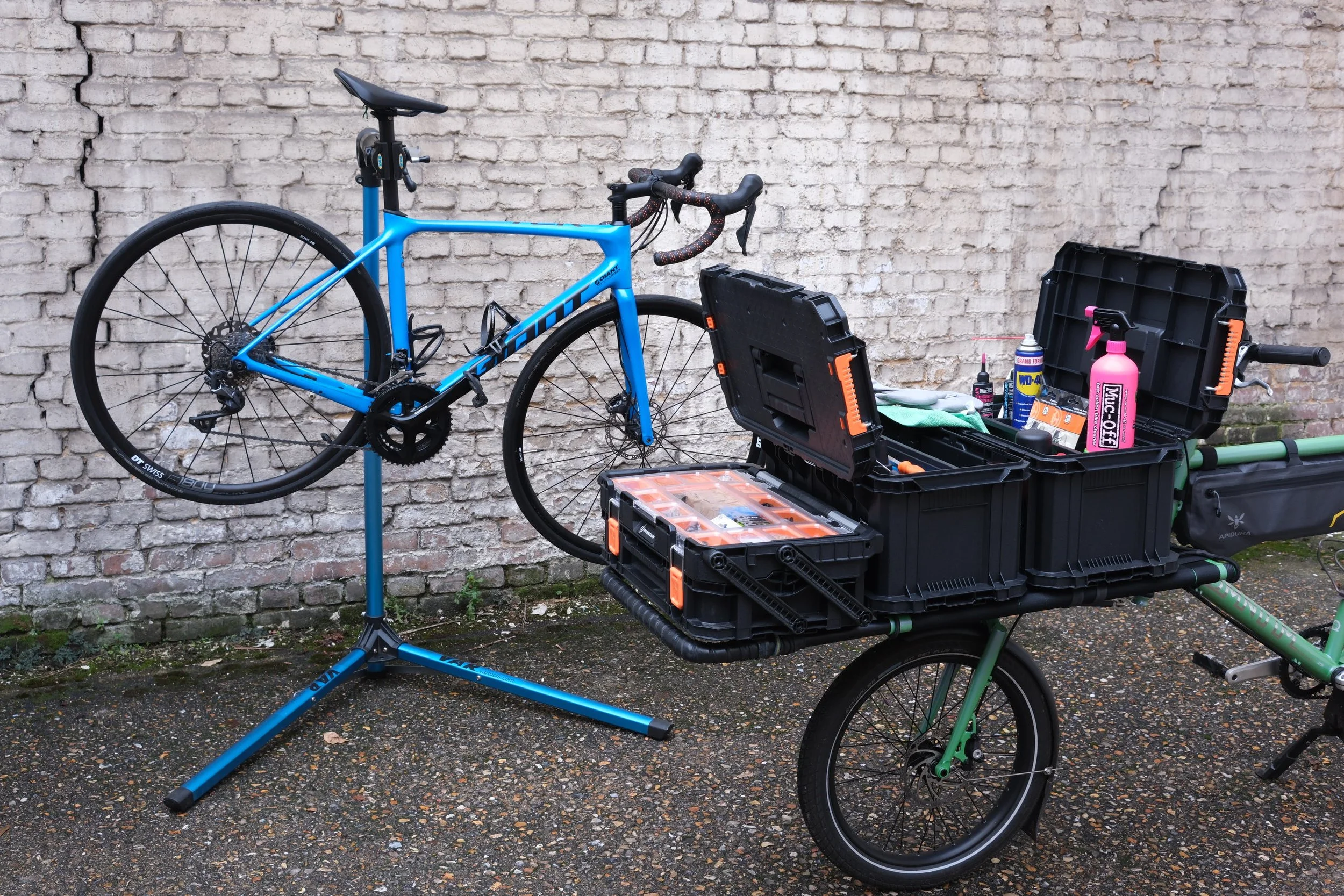 Un vélo fixé sur un support et une remorque contenant des outils et des produits d'entretien, contre un mur en briques.
