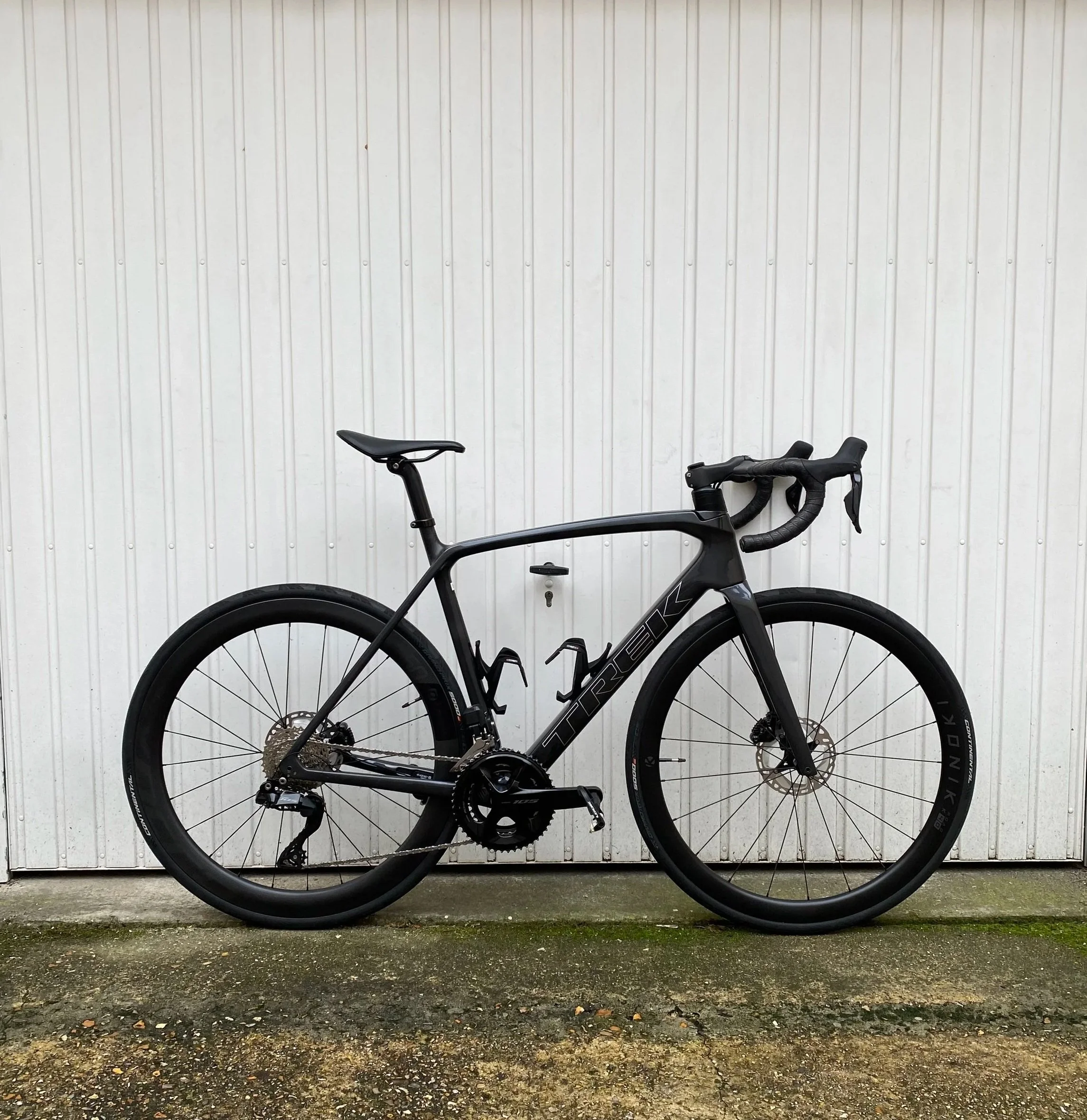 Vélo de route noir avec cadre carbone, pneus slick, et guidon en drops, stationné contre un mur en panneaux de métal blanc.