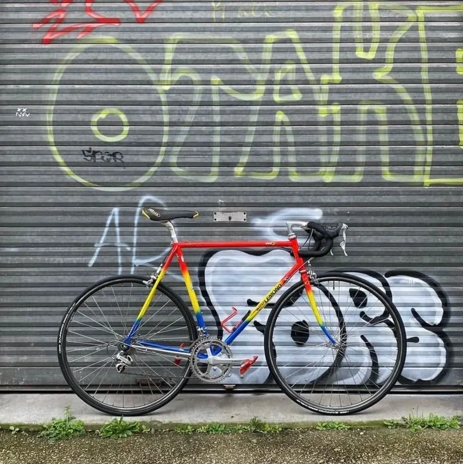 Un vélo rouge avec des bandeaux de couleurs vives est stationné devant un mur avec du graffiti en gris, jaune, blanc et noir.