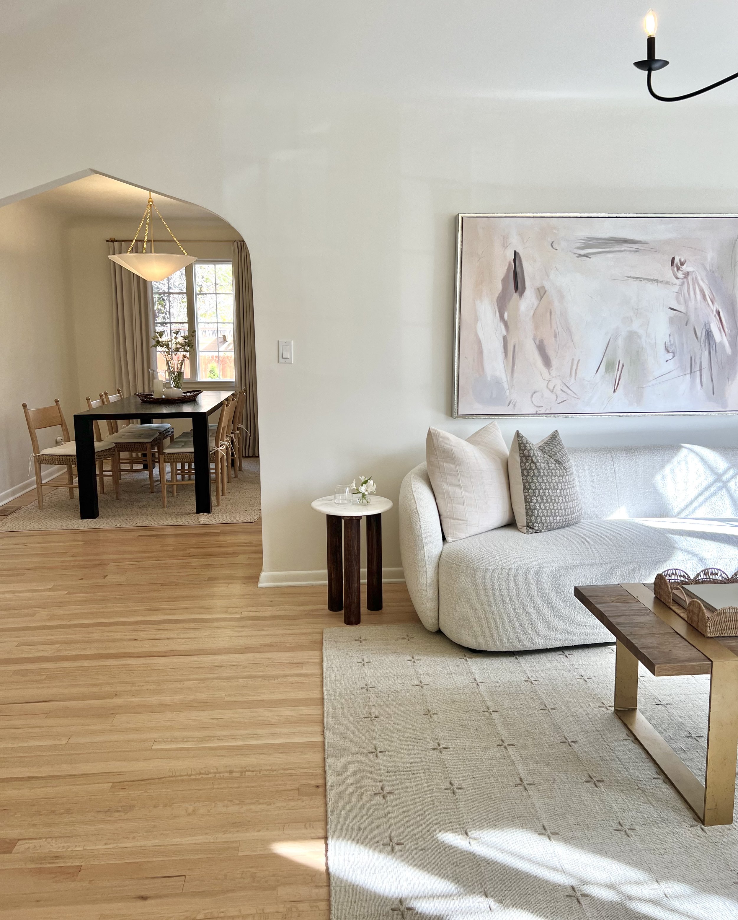 A living room with a white curved sofa, pillows, a wooden coffee table, a beige rug, and a large abstract painting on the wall. A small side table with flowers and glasses stands next to the sofa. Sunlight streams through a window, casting shadows.