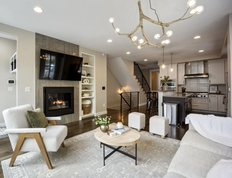 Living room with a modern fireplace, a wall-mounted TV, a white armchair, a round coffee table with books and a plant, a sofa, and an open kitchen with a central island, pendant lights, and stainless steel appliances.