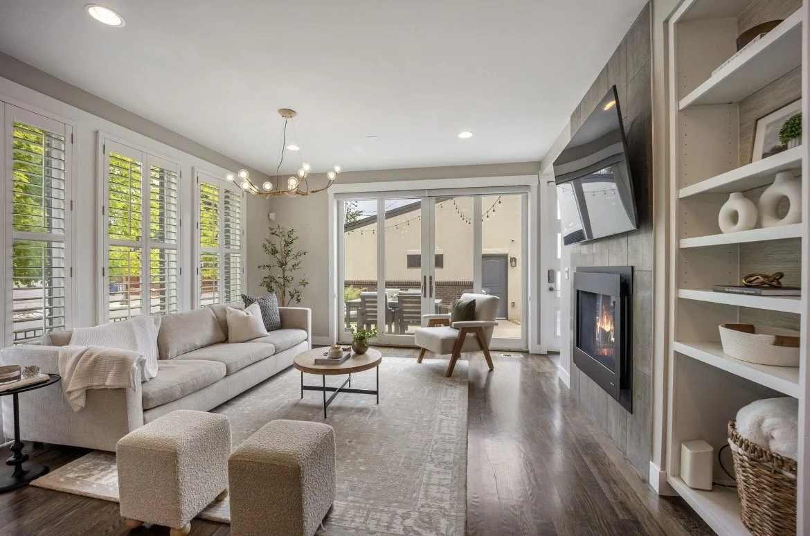 A living room with a white sectional sofa, a round coffee table, and ottomans. Large windows with white shutters, a sliding glass door leading outside, and a wall-mounted TV above a fireplace. Shelves with decorative items and a potted plant, with wood flooring and a beige rug.