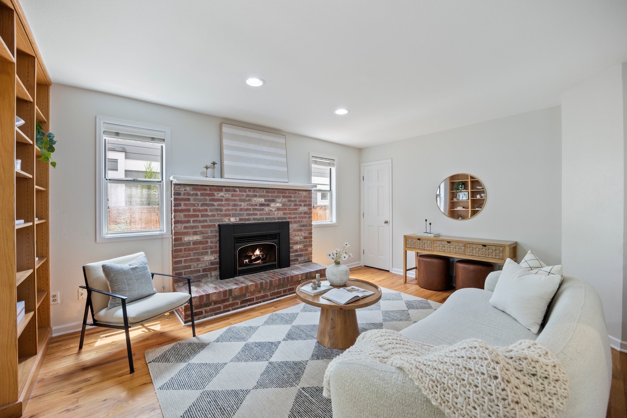Living room with a brick fireplace, white walls, and hardwood floors. Contains a white sofa with pillows, a round wooden coffee table with decor, a patterned rug, a white armchair with a pillow, a wooden console table with pillows, a round mirror, and built-in wooden shelves.