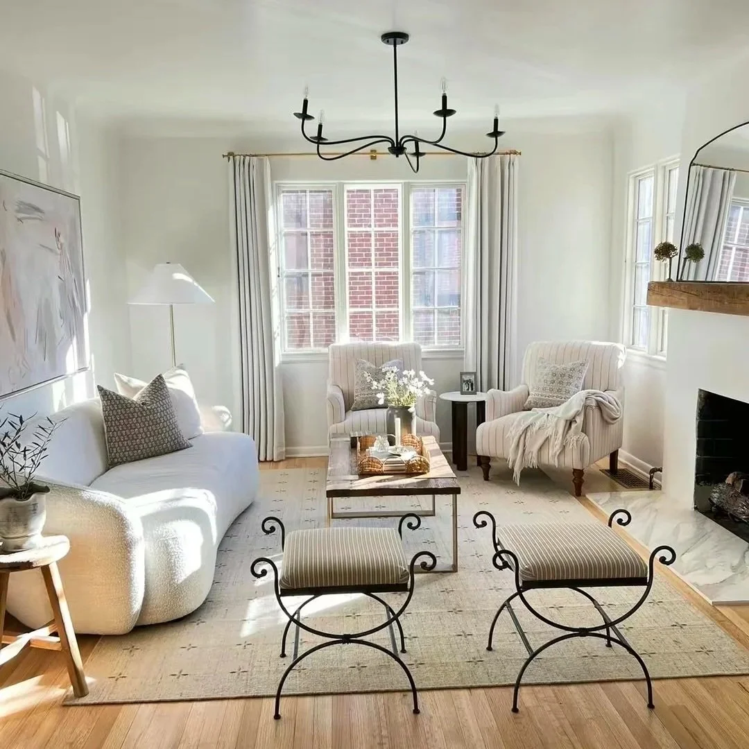 Bright living room with white sofa, armchairs, black chandelier, wooden coffee table, and large windows with white curtains, light wood flooring, fireplace, and minimal decor.
