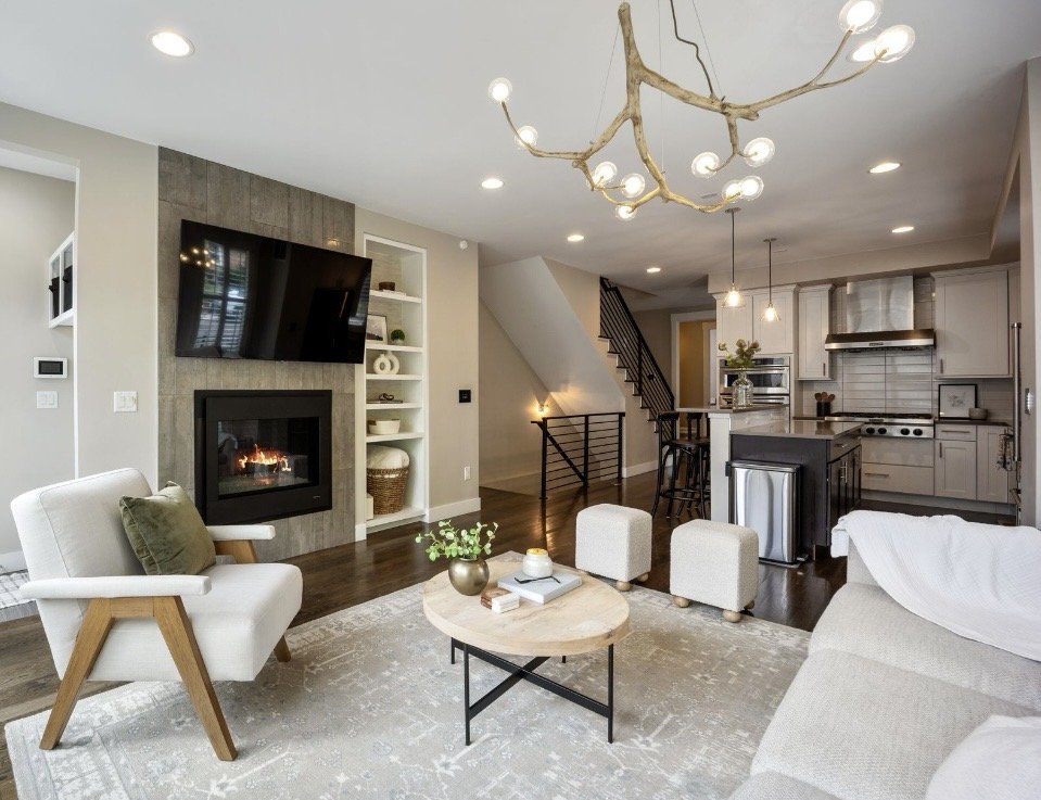 Modern living room with fireplace, wall-mounted TV, beige armchair with cushion, round coffee table with decor items, white sofa with blanket, kitchen with island, stainless steel appliances, and staircase with black railing, all decorated in neutral