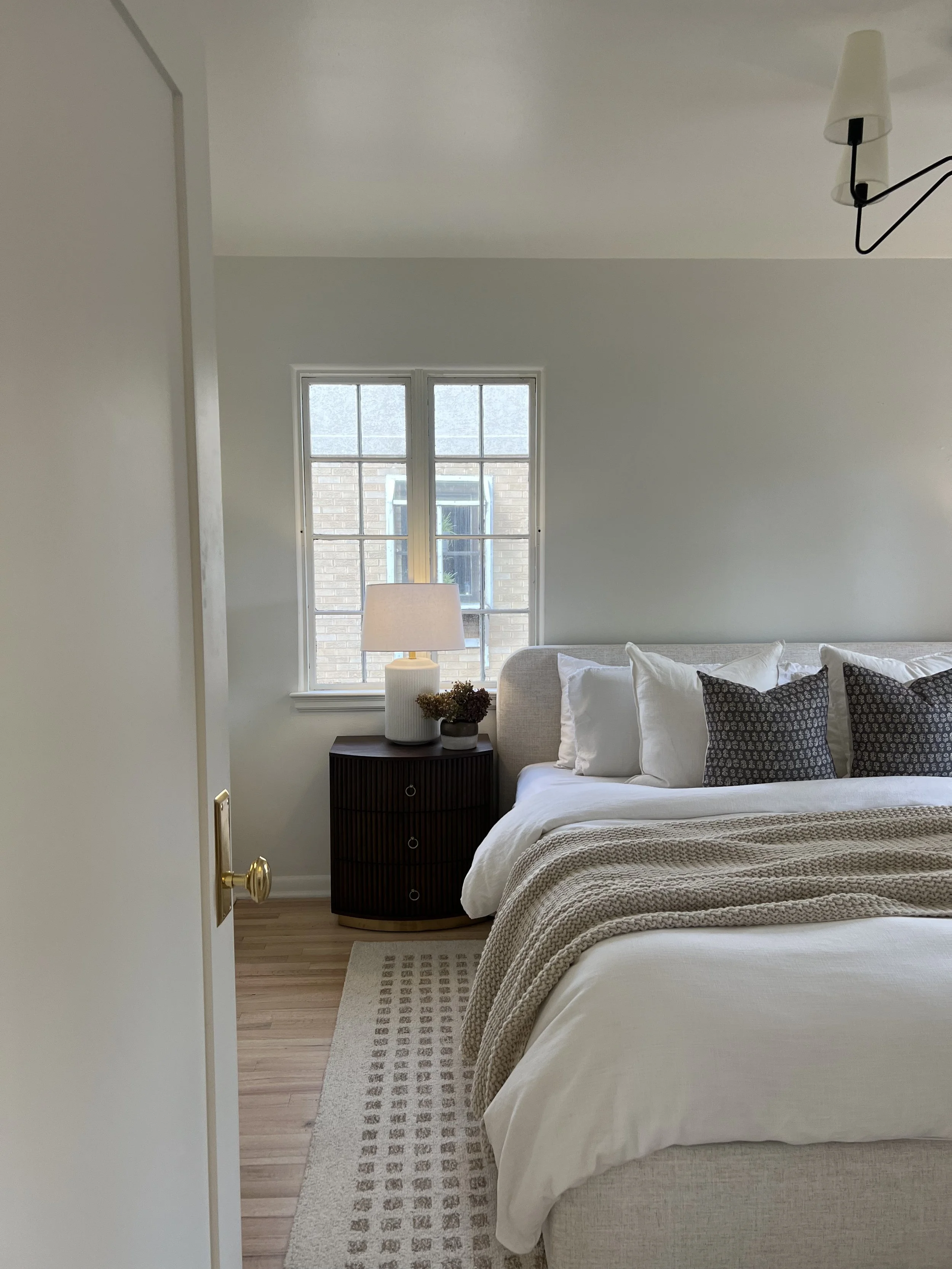 A bedroom with a bed, pillows, a beige blanket, a nightstand with a lamp, and a window with a view of a brick building.