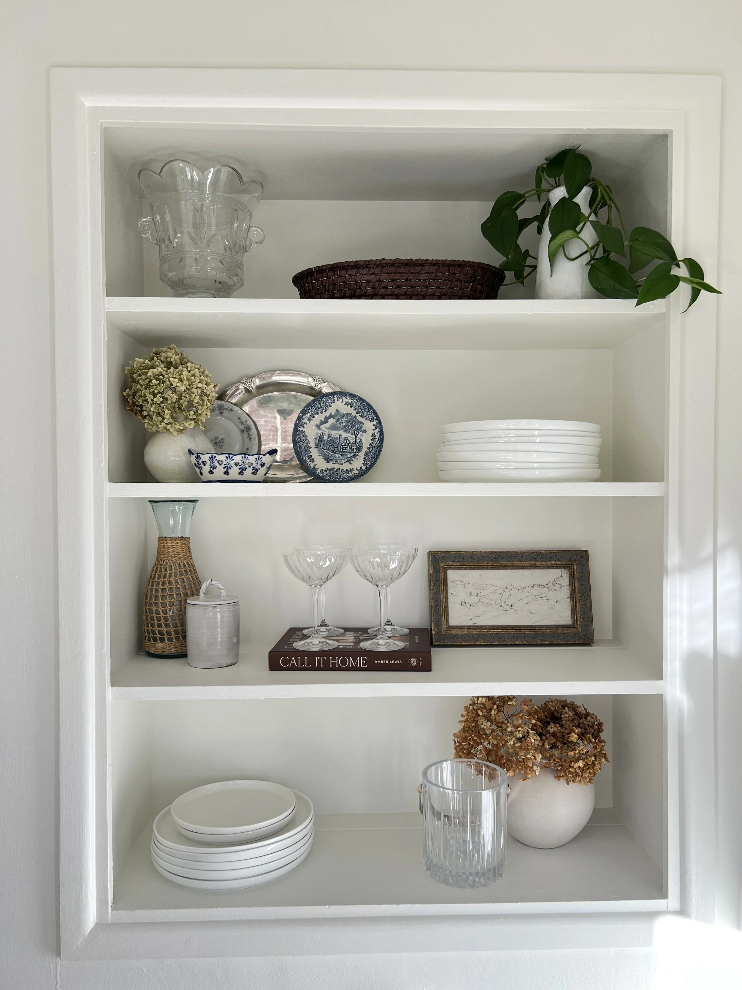 White built-in shelf with decorative dishes, bowls, a vase of dried flowers, a framed picture, glasses, and a potted plant, all arranged in a tidy display.