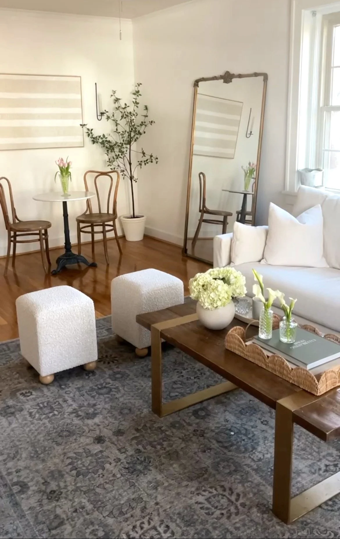 A cozy living room with a white sofa, a wooden coffee table with white flowers, a patterned rug, and two white ottomans. In the background, there is a small dining area with a round table, two wooden chairs, a large potted plant, a tall mirror, and a wall art piece.