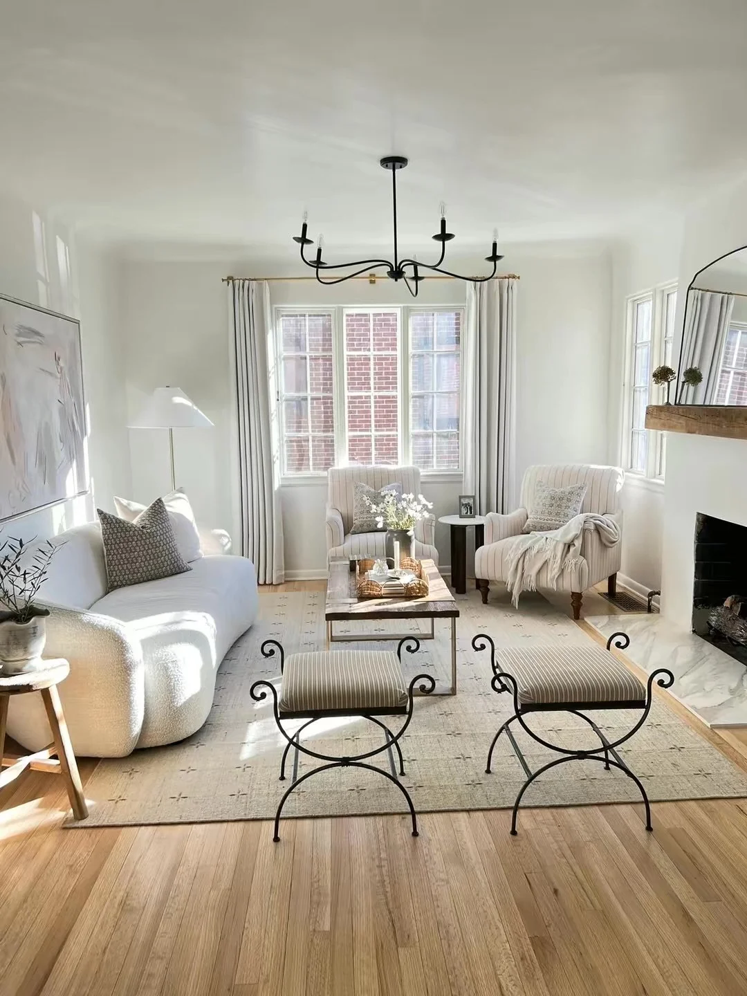 Bright living room with white walls, wooden floor, and large windows. Contains white sofa, striped armchairs, a wooden coffee table, and two metal-framed stools. Decor includes a chandelier, artwork, potted plants, and cushions.