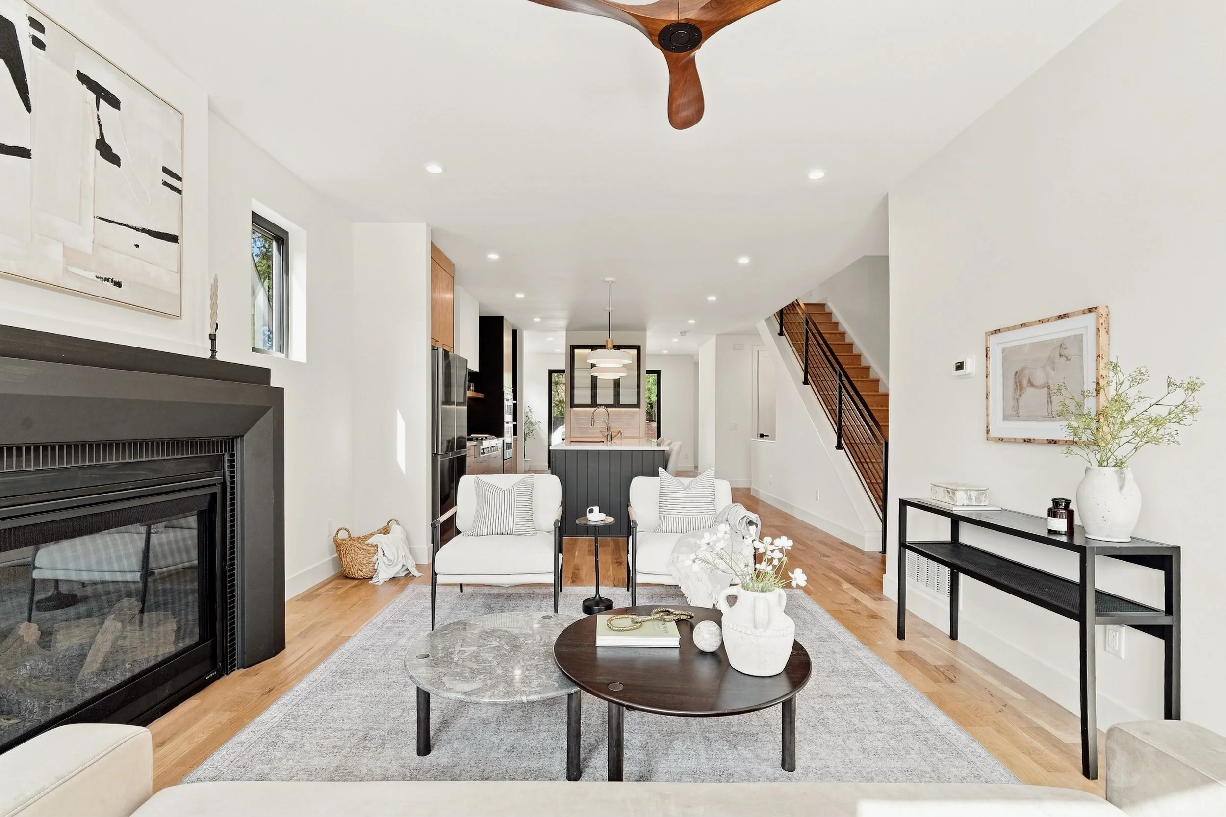 A living room with a white sectional sofa, a round coffee table, and ottomans. Large windows with white shutters, a sliding glass door leading outside, and a wall-mounted TV above a fireplace. Shelves with decorative items and a potted plant, with wood flooring and a beige rug.