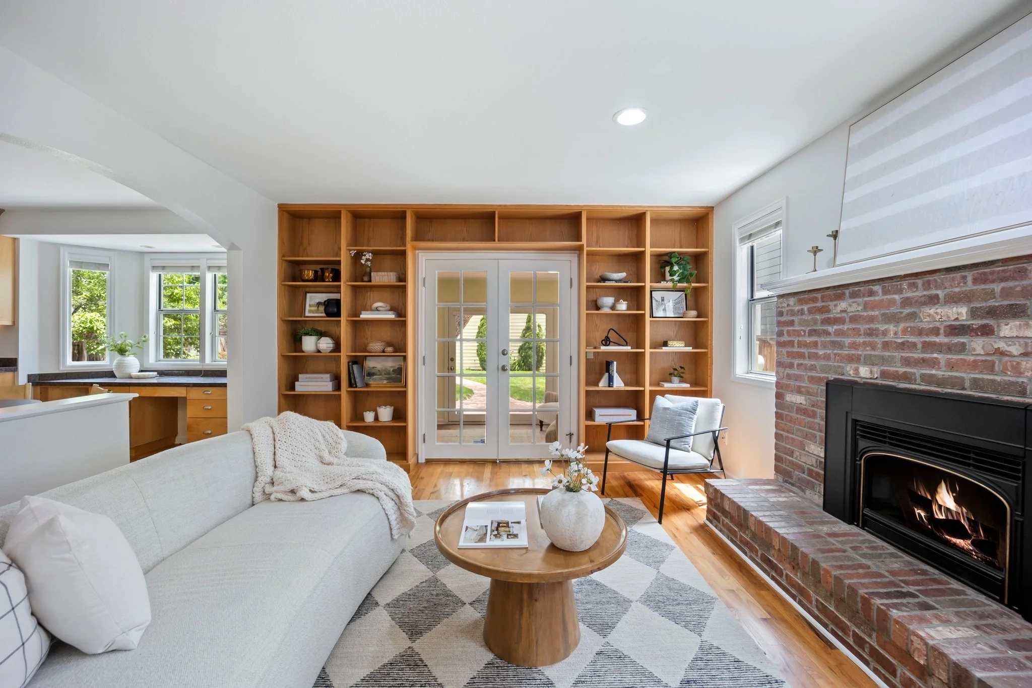 Living room with a white sofa, a wooden coffee table with a white vase and flowers, a brick fireplace with a fire, built-in wooden shelves, and French doors leading outside