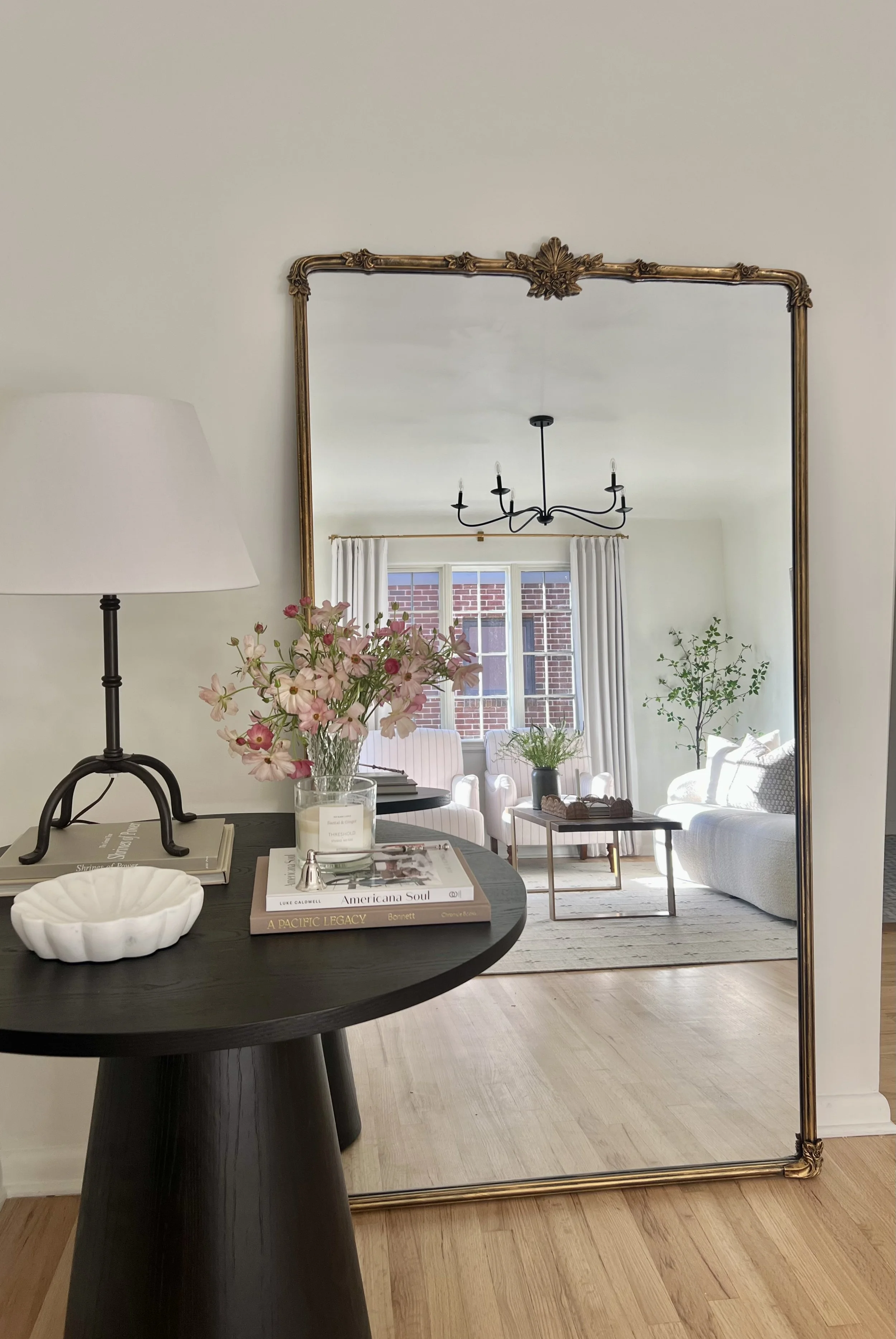 A large ornate mirror reflecting a bright living room with white sofas, a black chandelier, and large windows with white curtains.