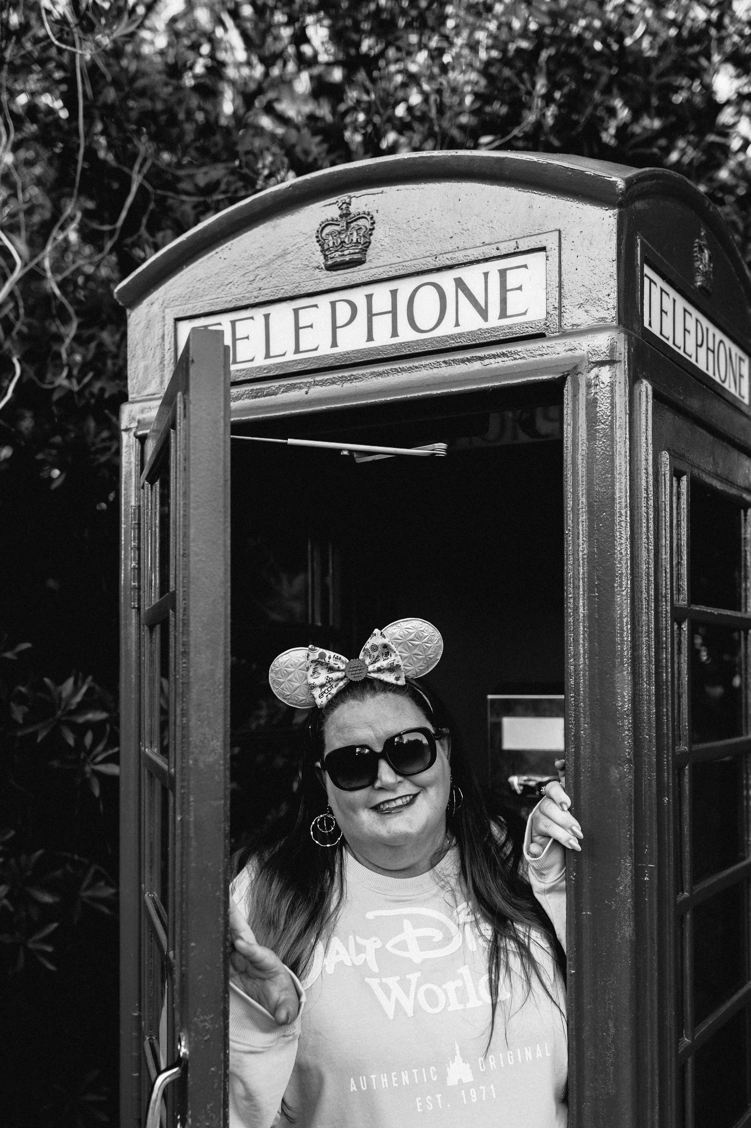 A woman standing inside a classic British telephone booth, smiling, wearing large sunglasses, Mickey Mouse ears headband, and a Walt Disney World sweatshirt.