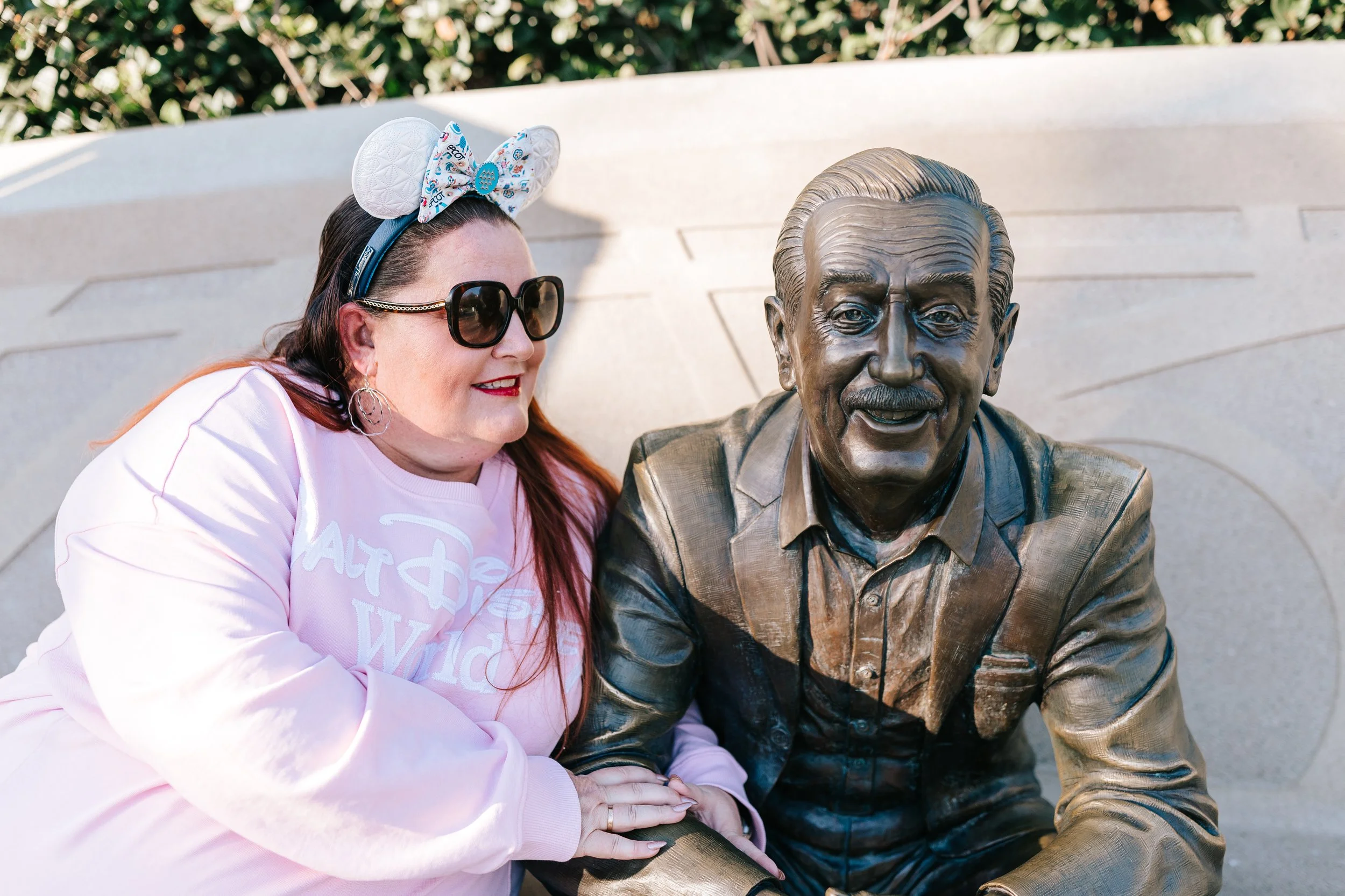A woman with long red hair, wearing sunglasses, a pink Walt Disney World sweatshirt, and Minnie Mouse ears headband, sitting next to a bronze statue of Walt Disney, who is smiling and wearing a suit.