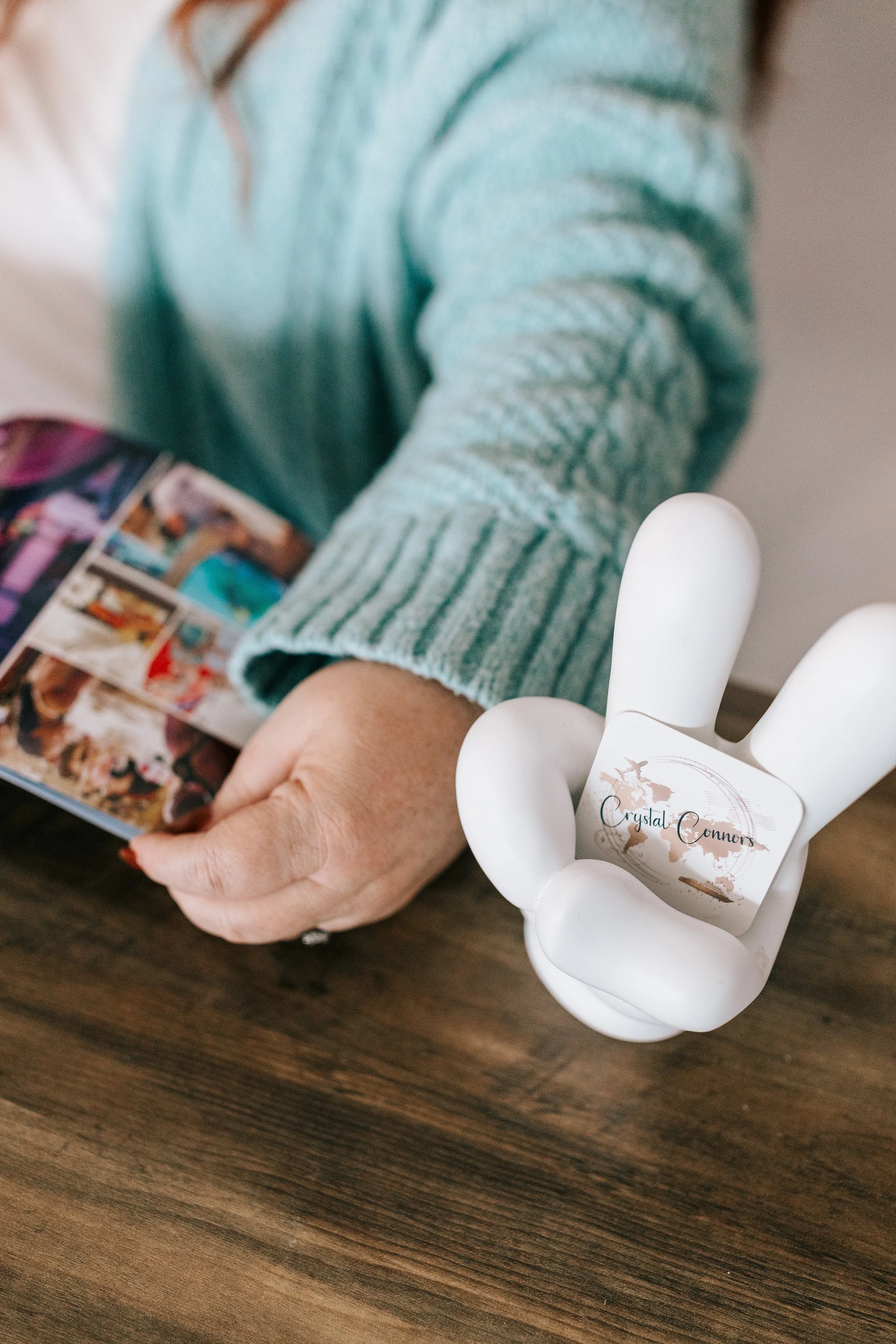 A woman in a green sweater holding a photo album, with a white ceramic hand-shaped business card holder displaying a card for Crystal Connors on a wooden table.