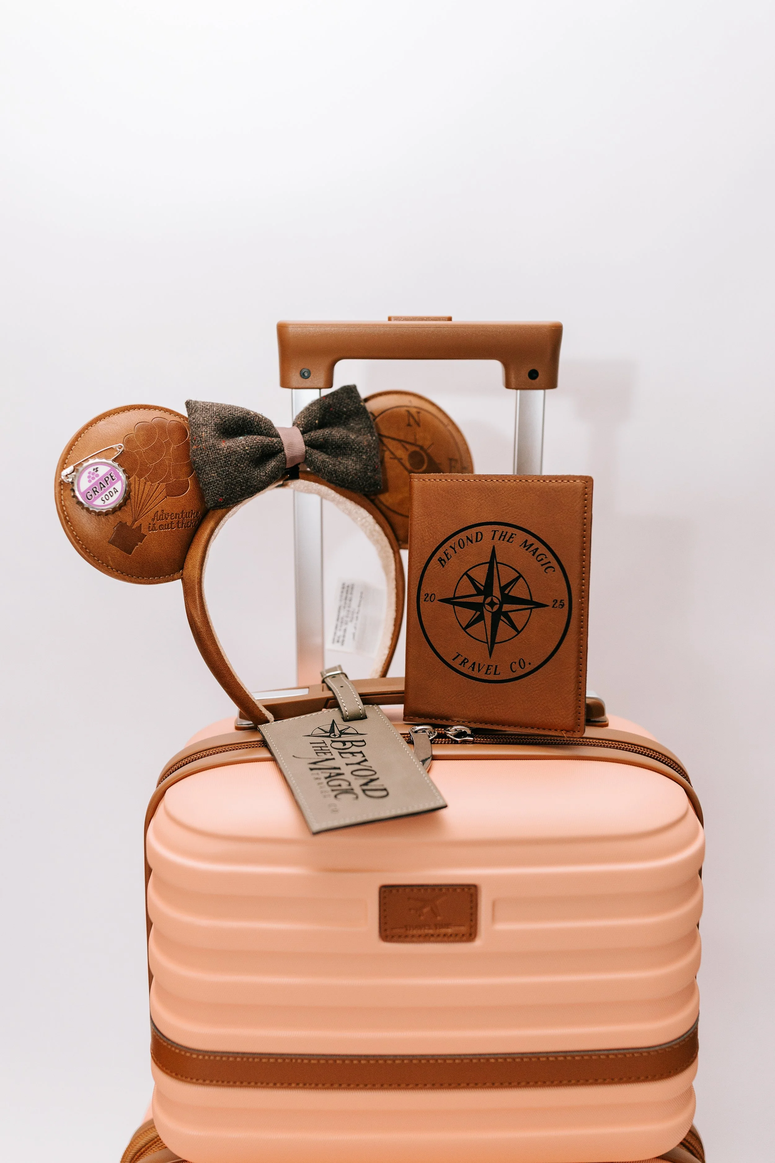 Pink suitcase with brown travel accessories on top, including a travel journal, a bow tie headband, and a travel-sized pouch, all against a plain white background.