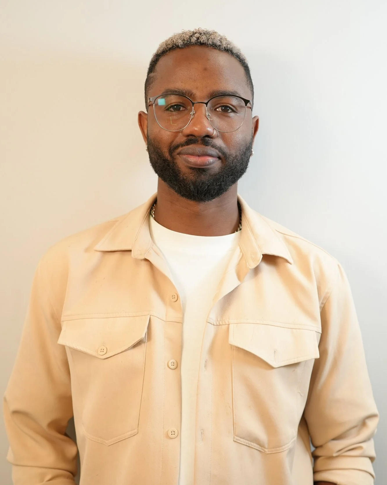 Portrait of a man with glasses, a beard, and short, light-colored hair, wearing a beige jacket over a white shirt, standing against a plain light-colored wall.