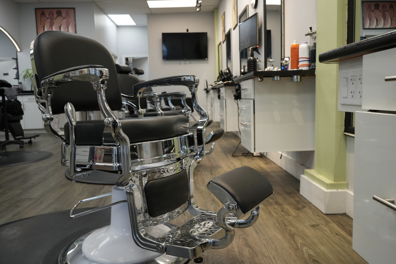Salon chair in a barber or hair salon, with hair styling tools and products on nearby counters, and a wall-mounted TV in the background.