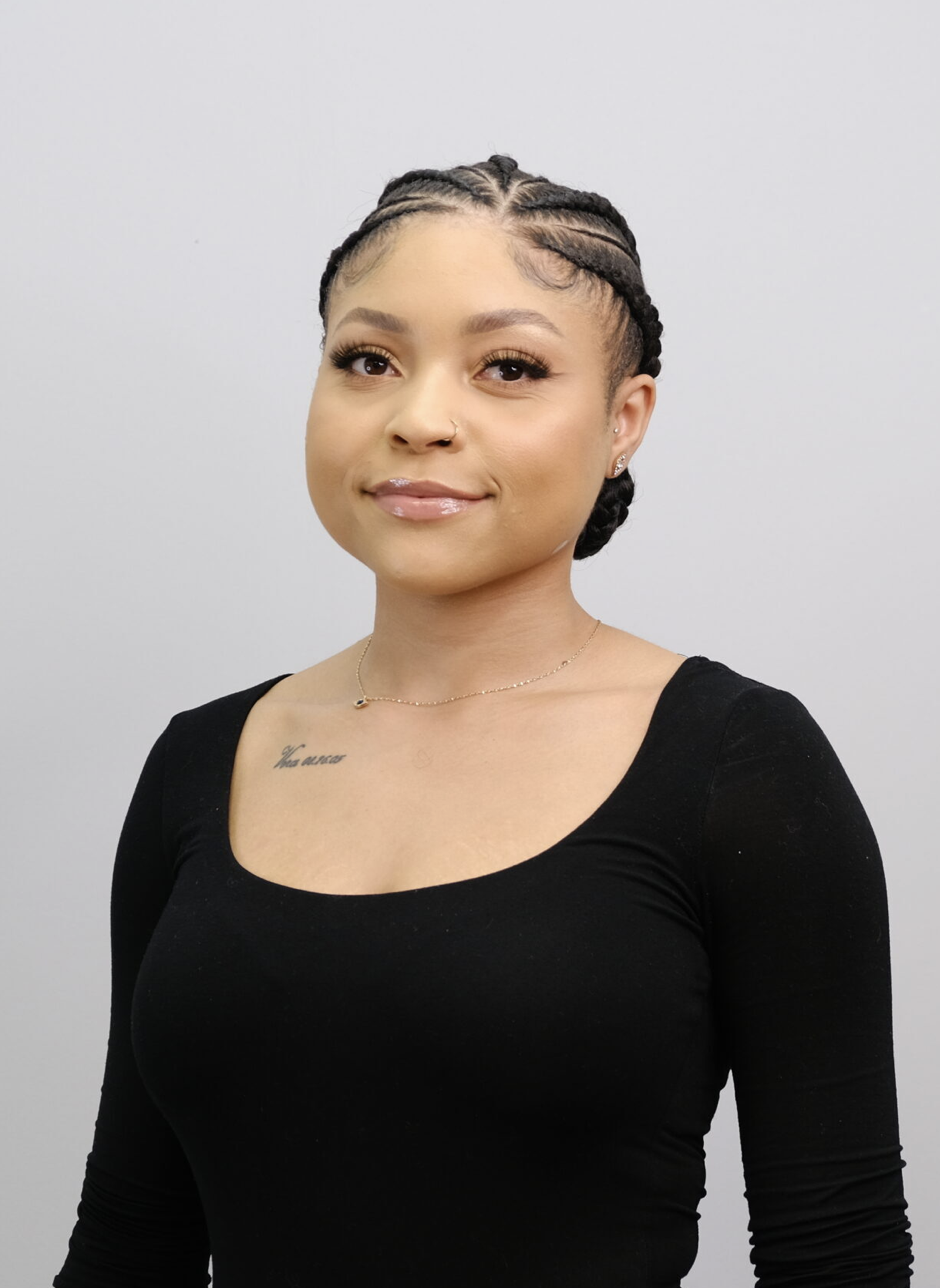A young woman with braided hair, wearing a black top and jewelry, standing against a plain background.