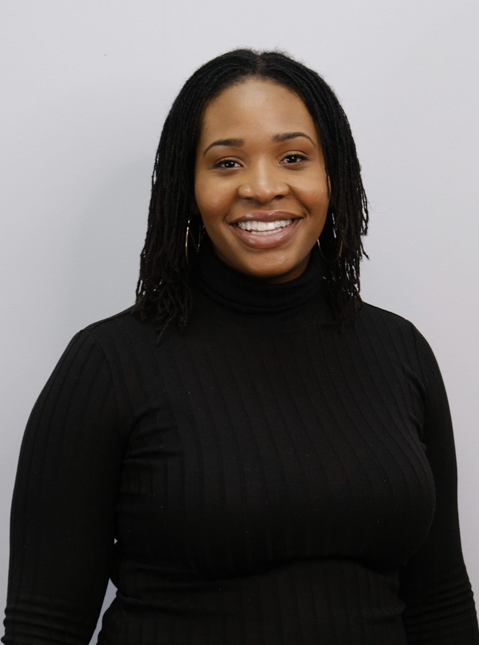 A woman with shoulder-length dreadlocks wearing a black turtleneck sweater, smiling, against a plain gray background.