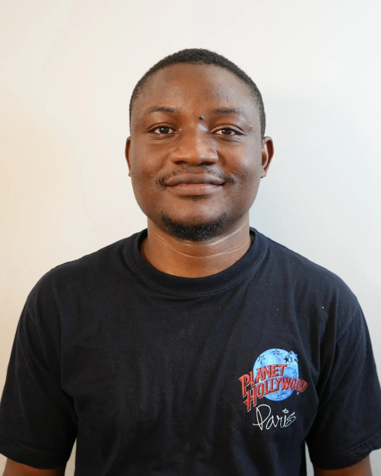 A young man with short hair and facial hair, wearing a black t-shirt with 'Planet Hollywood Paris' logo, standing against a plain white wall.
