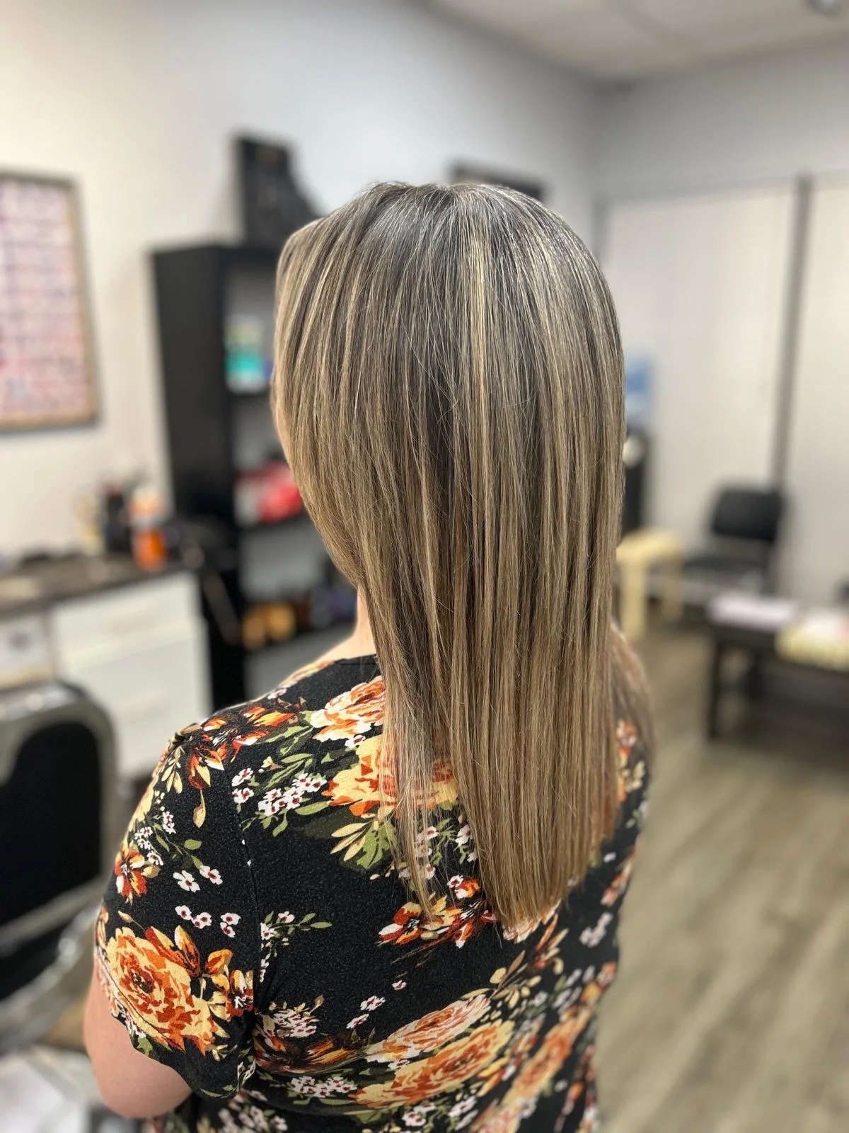Back view of a woman with long, straight, shoulder-length blonde hair with highlights, wearing a black floral dress.