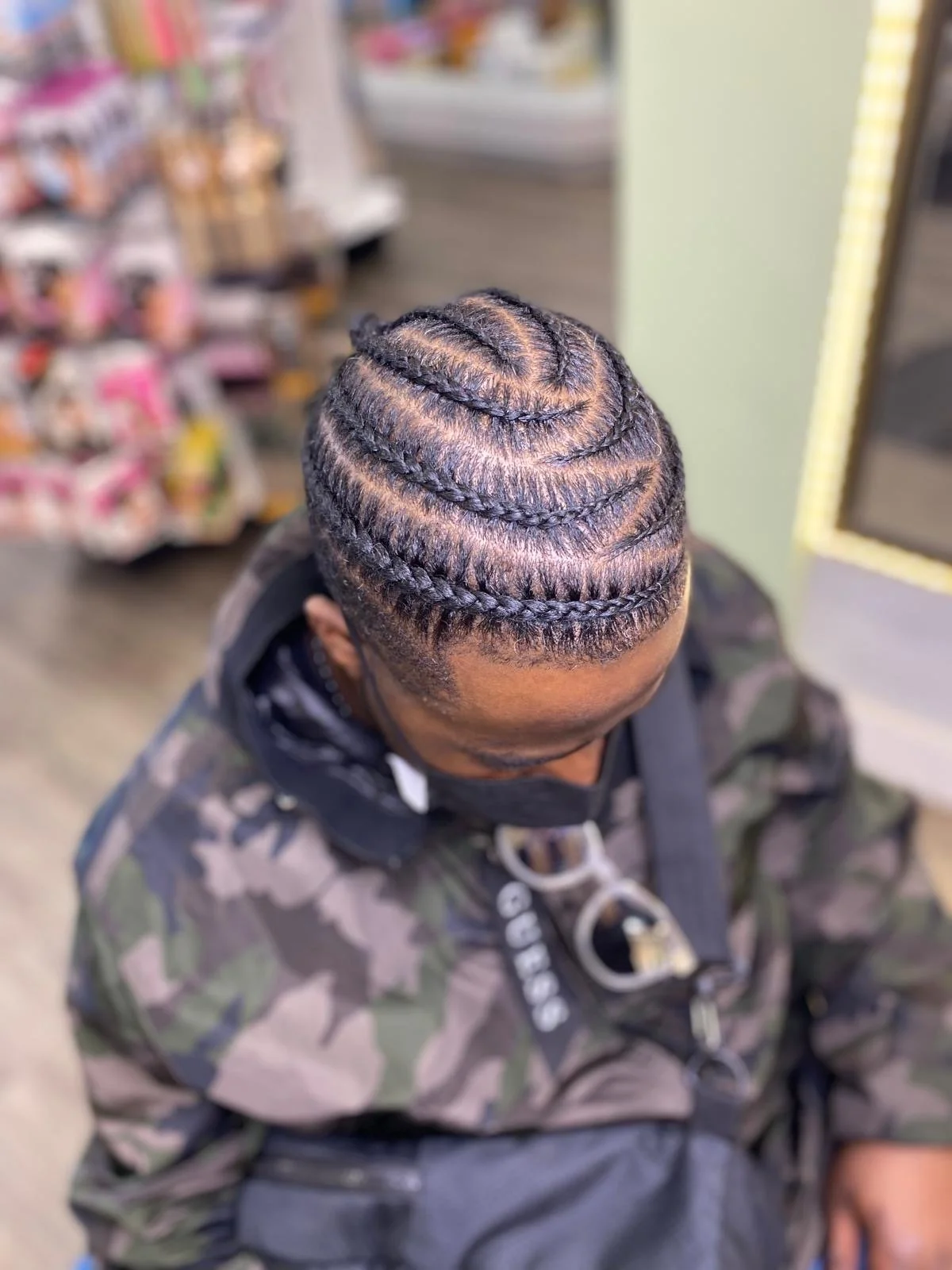 A person with intricately braided cornrows in a spiral pattern, sitting indoors, wearing a camouflage jacket.