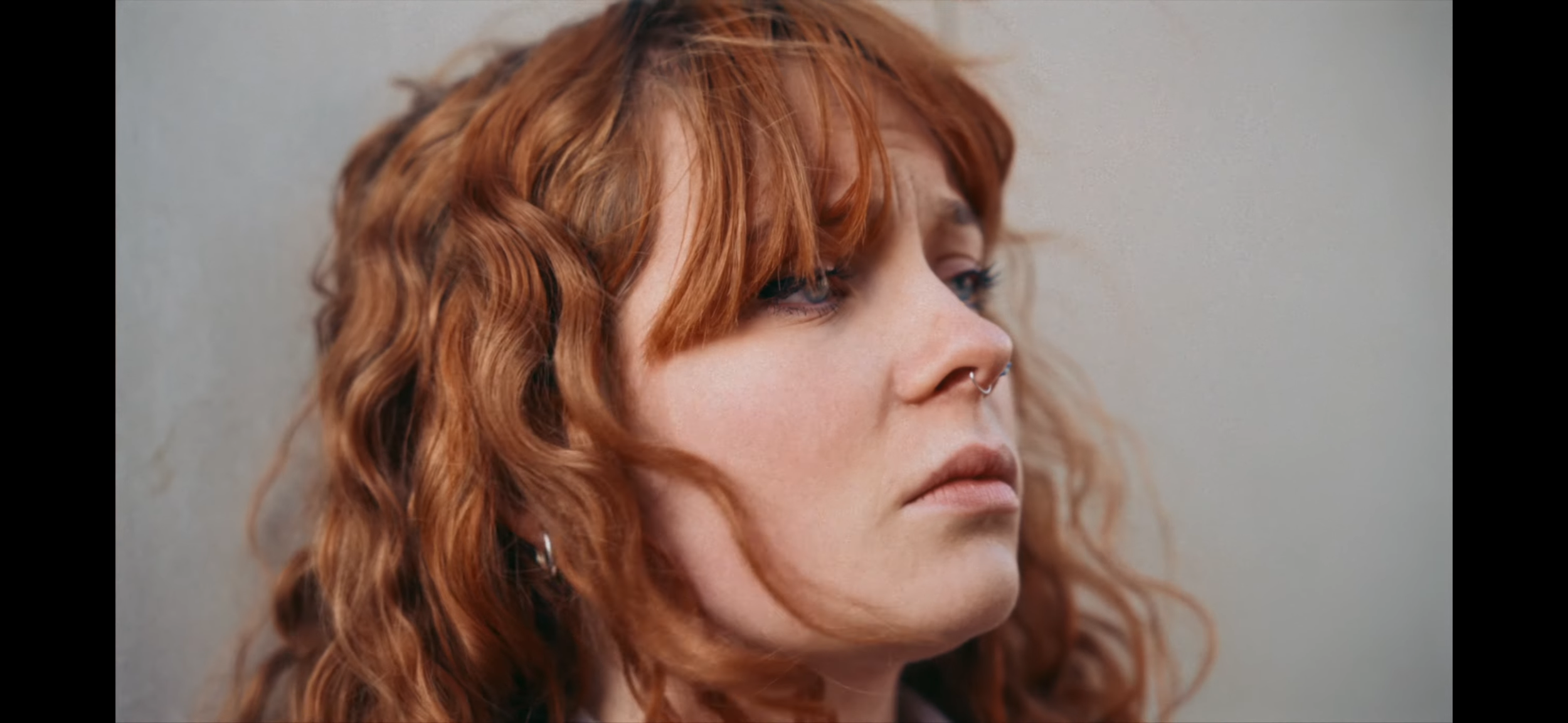 Close-up of a woman with red, curly hair, light skin, blue eyes, and a septum piercing, looking to the right against a plain background.