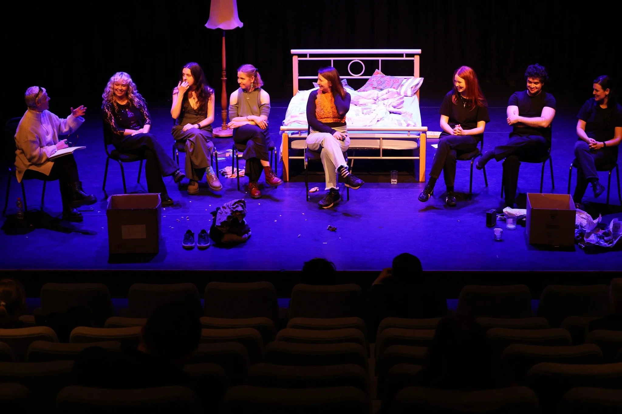 A stage production with nine actors sitting in a row, engaged in a discussion or performance. The set includes a bed and a lampshade, with props and clutter scattered on the stage floor.
