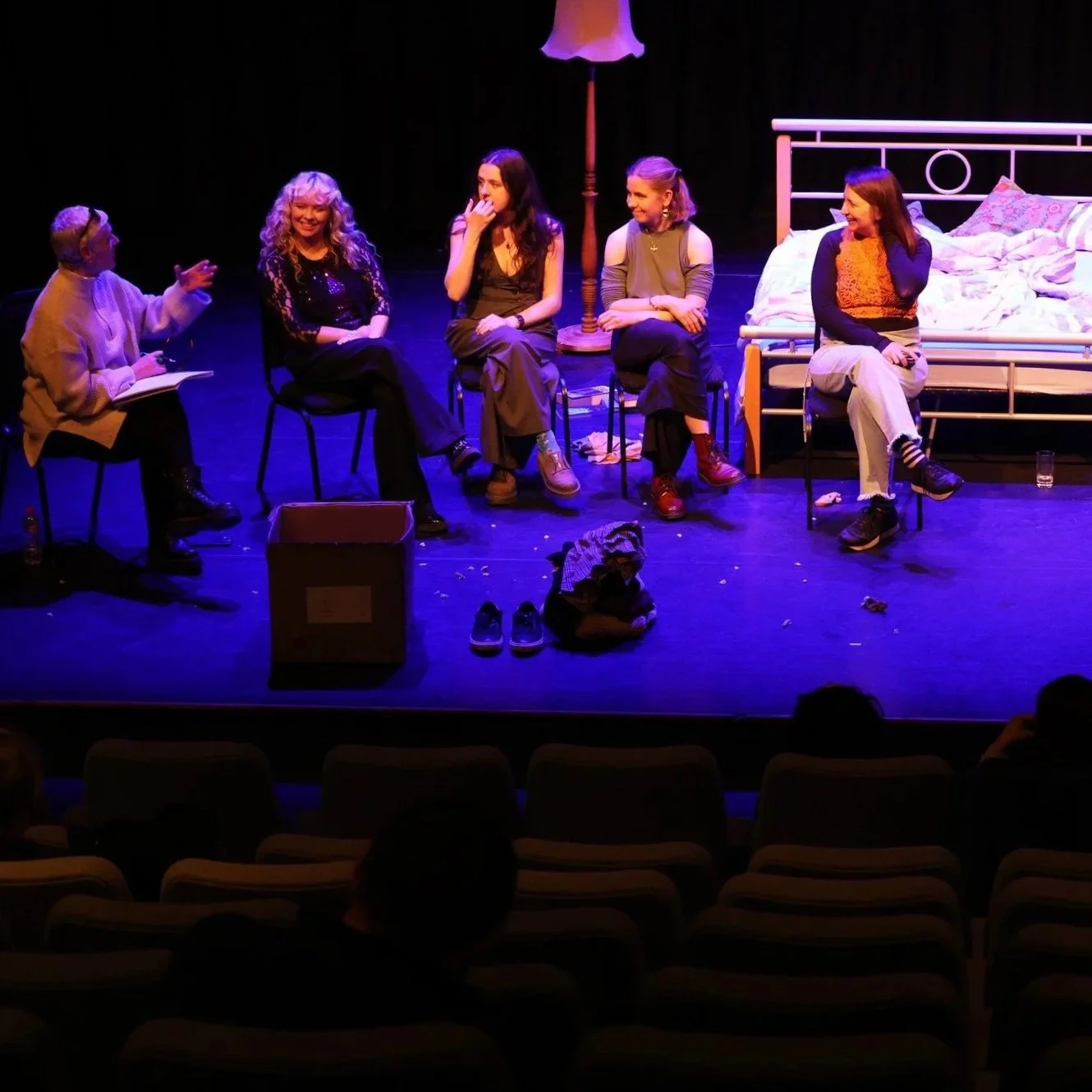 Stage scene with five women sitting on chairs, engaged in conversation, with a bed in the background and various items scattered on the floor.