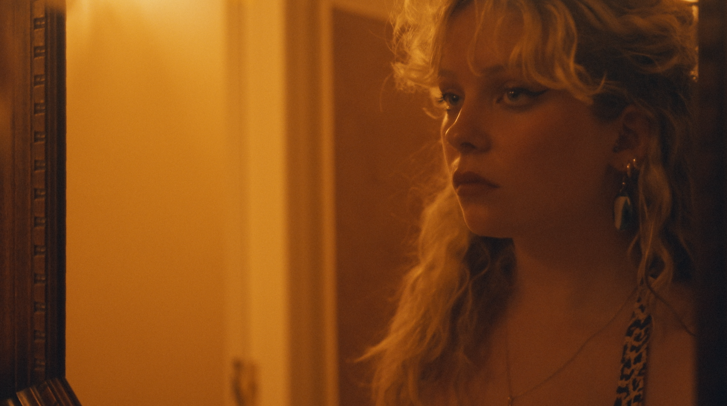 A woman with curly blonde hair and earrings looking to the side in a warm, dimly lit room.