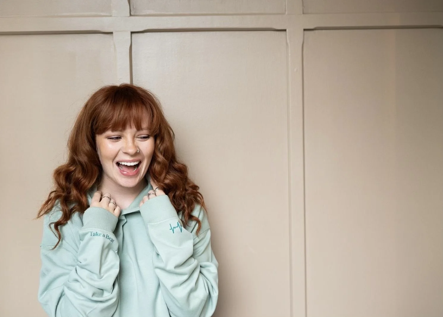 A woman with red hair and a nose ring smiling and holding her hands near her chest, standing in front of beige cabinets, wearing a light blue hoodie that says 'Take a Bear'.