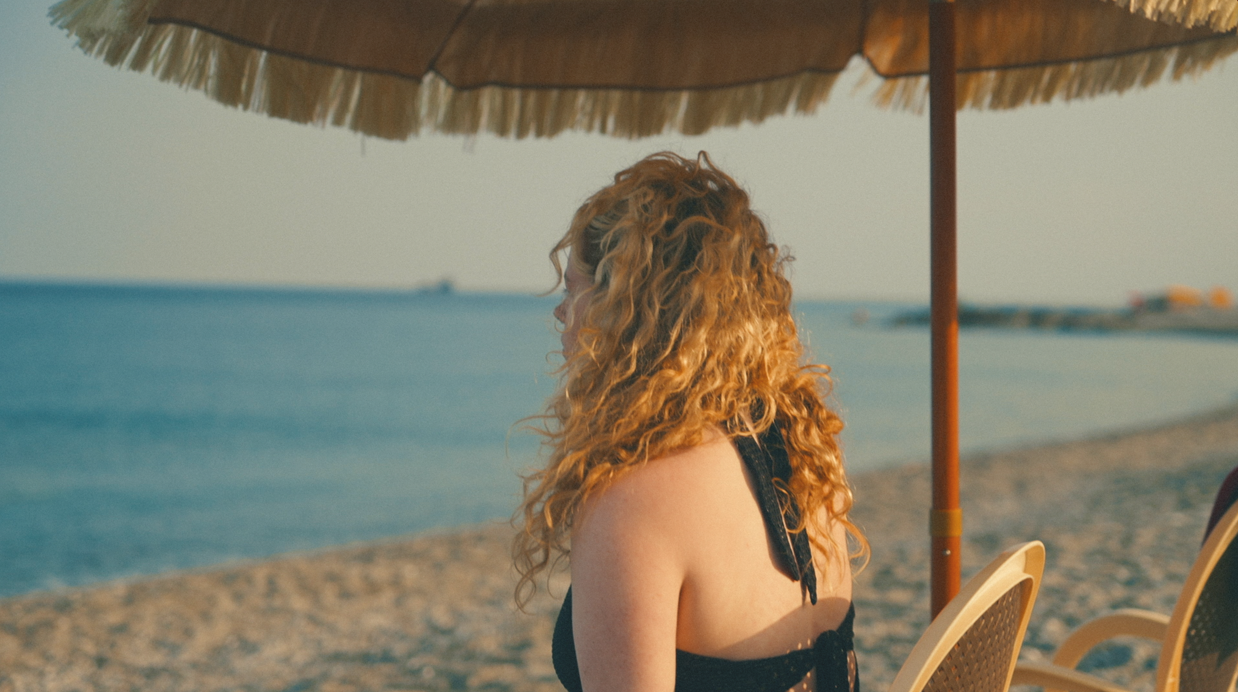 A woman with long curly red hair sitting under a beach umbrella on the sand, facing away from the camera, near the sea.