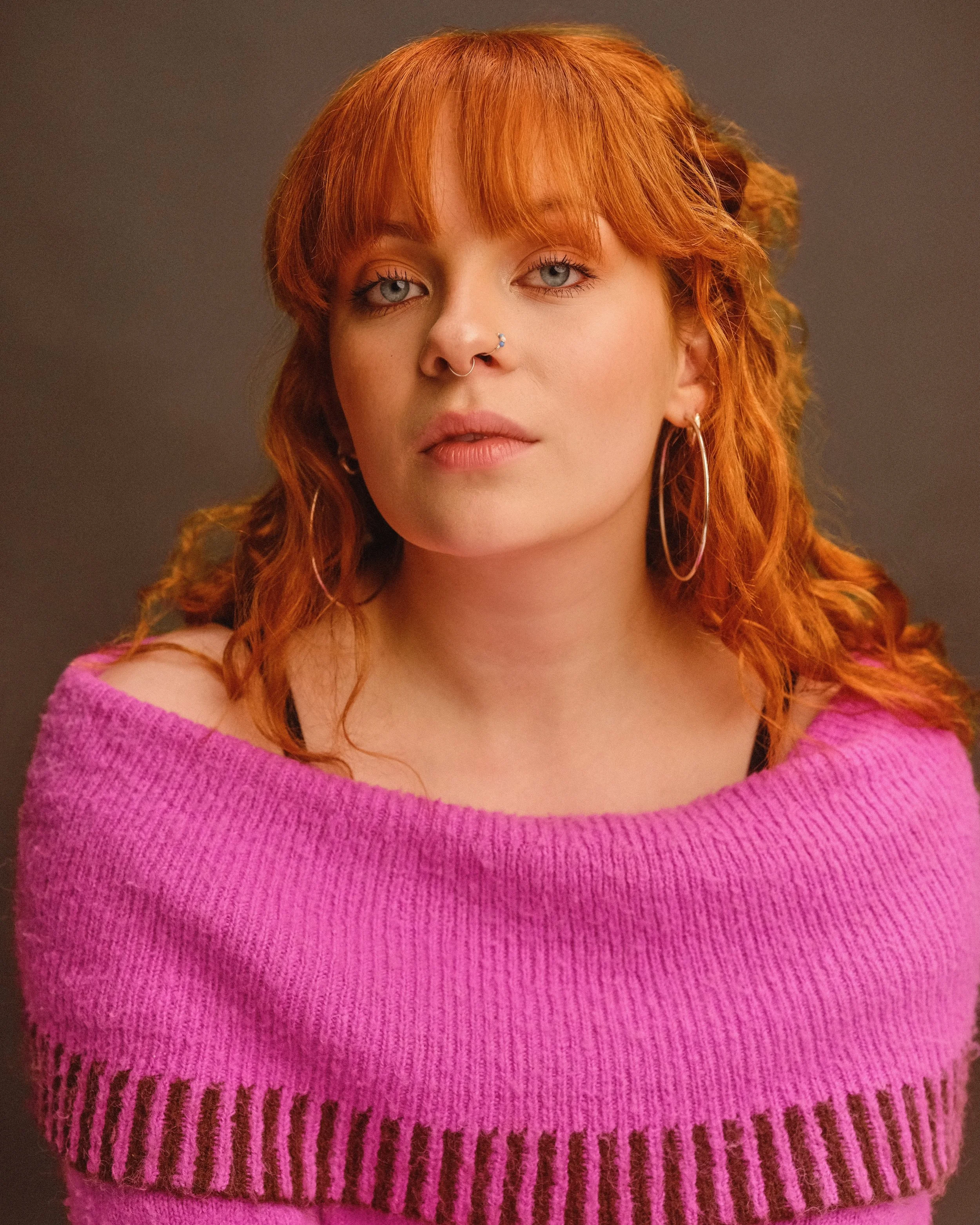 Close-up of a woman with red wavy hair wearing a pink sweater with open-knit pattern details at the bottom and large hoop earrings.