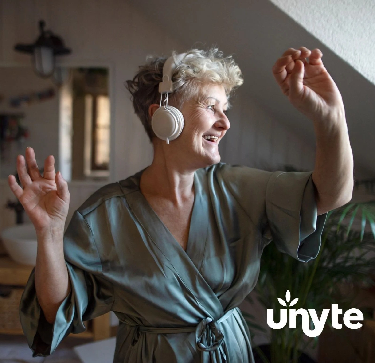 A smiling older woman with short gray hair wearing large white headphones, dancing and raising her arm in a cozy indoor space with a window and plants in the background.