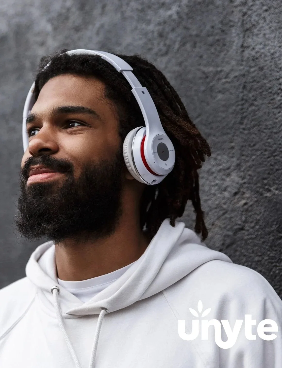 A man with dreadlocks and a beard wearing white headphones and a white hoodie, standing against a textured black wall.