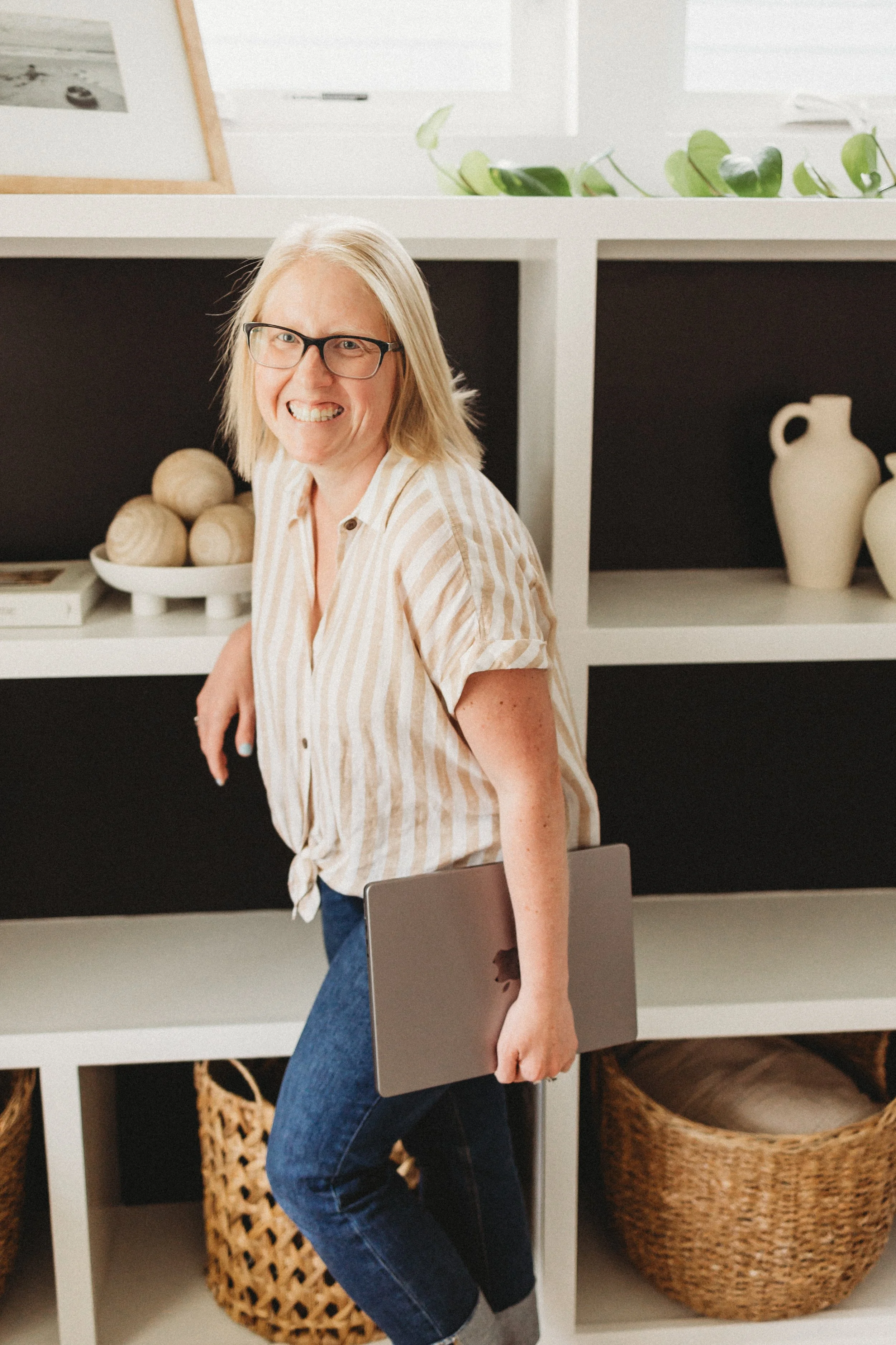 A smiling woman with glasses and blonde hair holding a closed laptop, standing in front of a white shelving unit with decorative items and baskets.