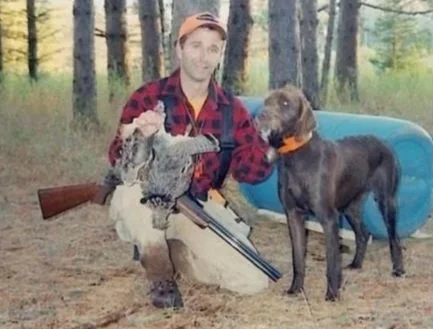 Man kneeling with a rifle and a hunting shotgun, holding a grouse, surrounded by a forest with a hunting dog.
