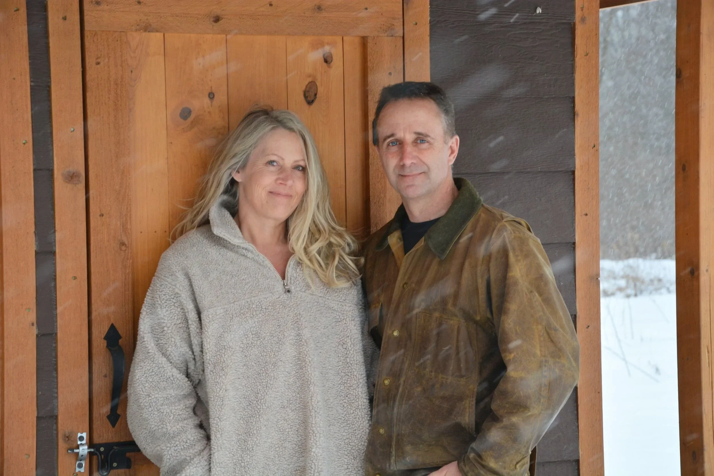 A with long blonde hair, wearing a light gray fleece jacket, standing next to a man with short dark hair, wearing a brown leather jacket, standing in front of a wooden door in a snowy outdoor setting.