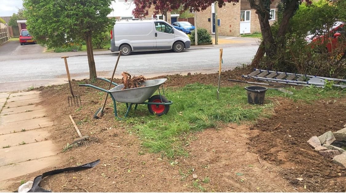 Garden clearance in Chepstow and Bristol. Garden area under renovation with tools, a wheelbarrow filled with dirt, rake, shovel, and ladder placed on the ground, and a street and houses in the background.