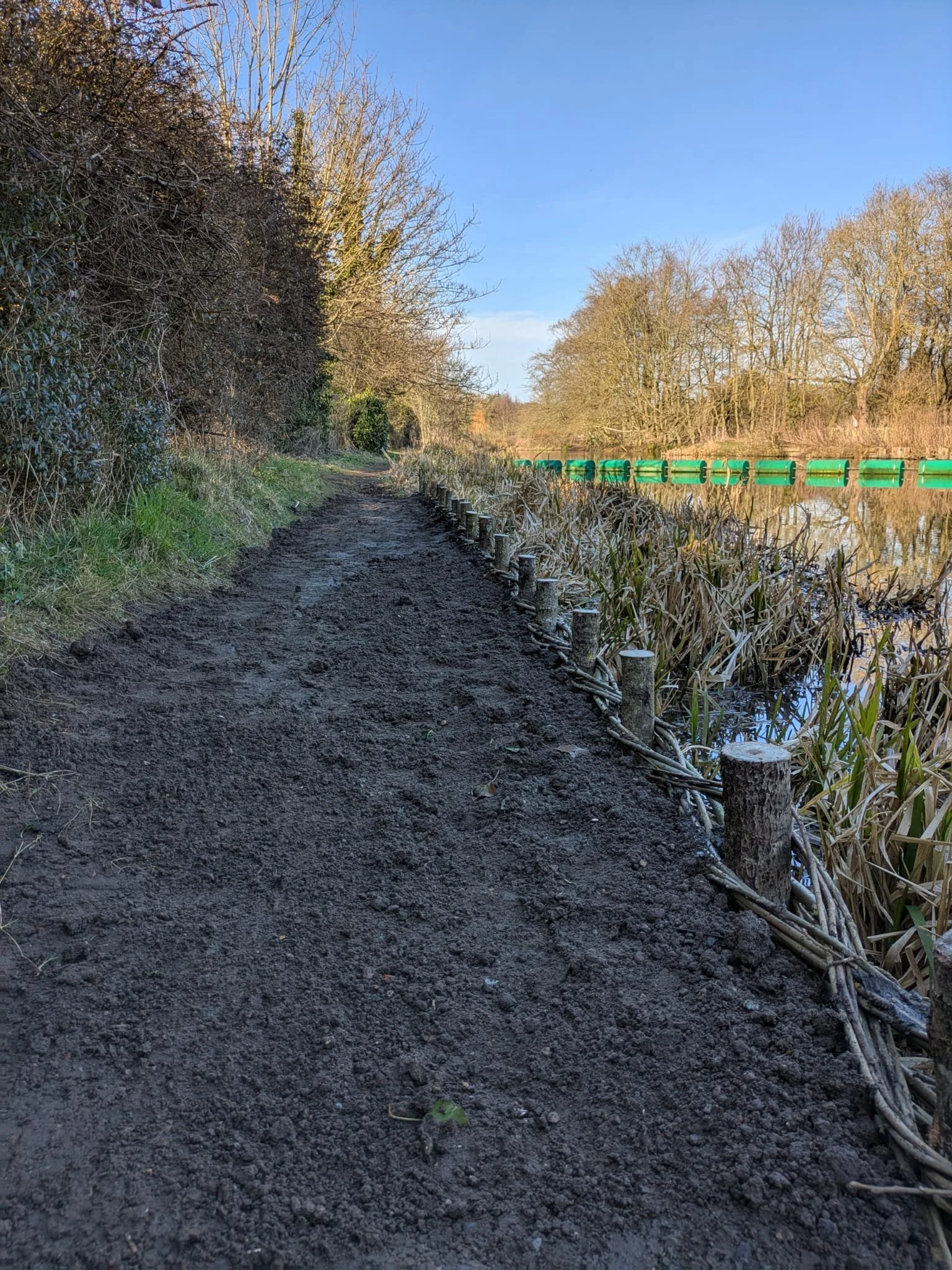 A dirt pathway along a water body with trees on both sides, a green barrier in the water, and clear blue sky.