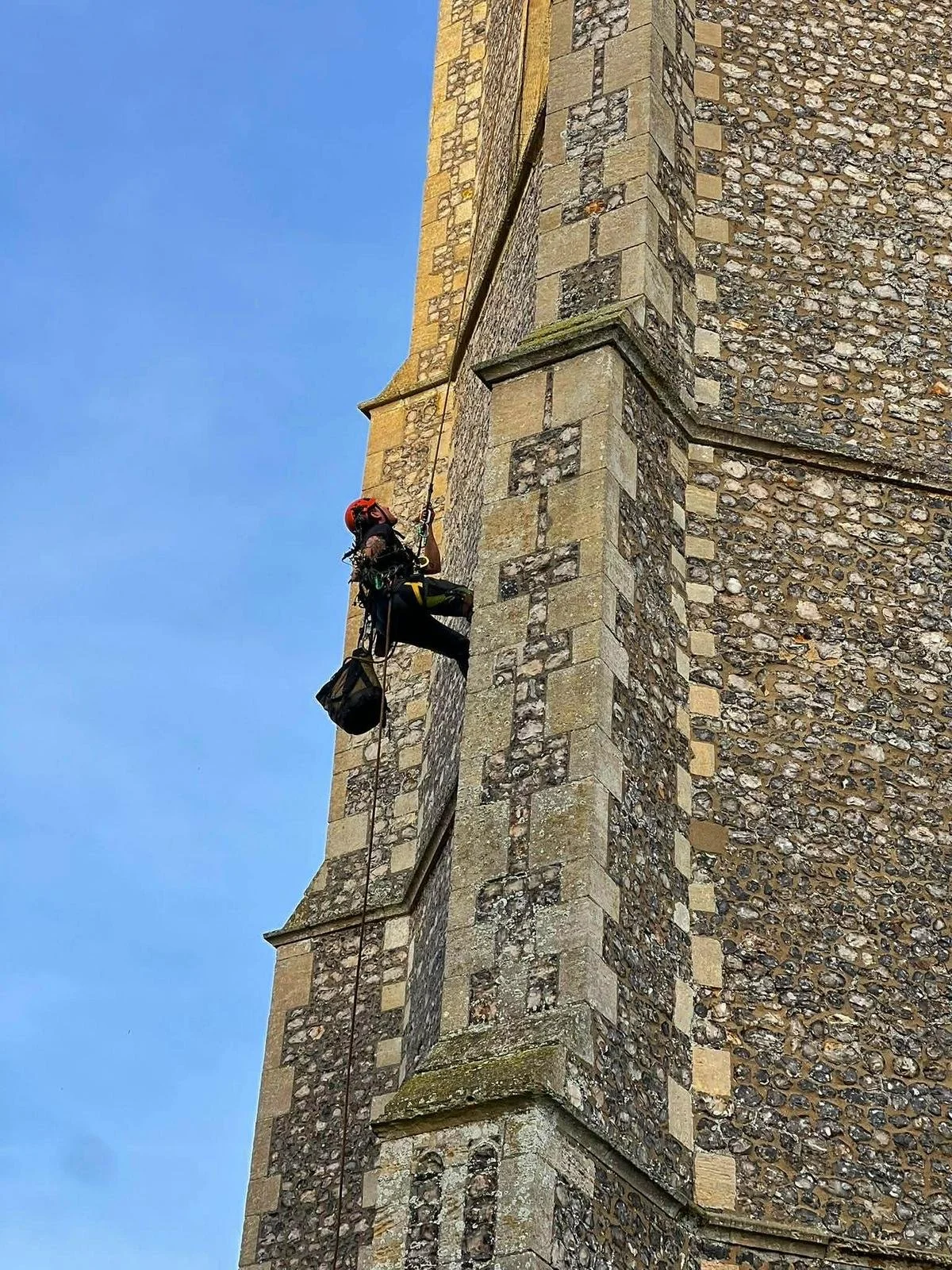 A person in climbing gear, including a helmet, harness, and ropes, is cleaning or inspecting the exterior of a tall stone building, possibly a church or historical structure, against a background of a clear blue sky.