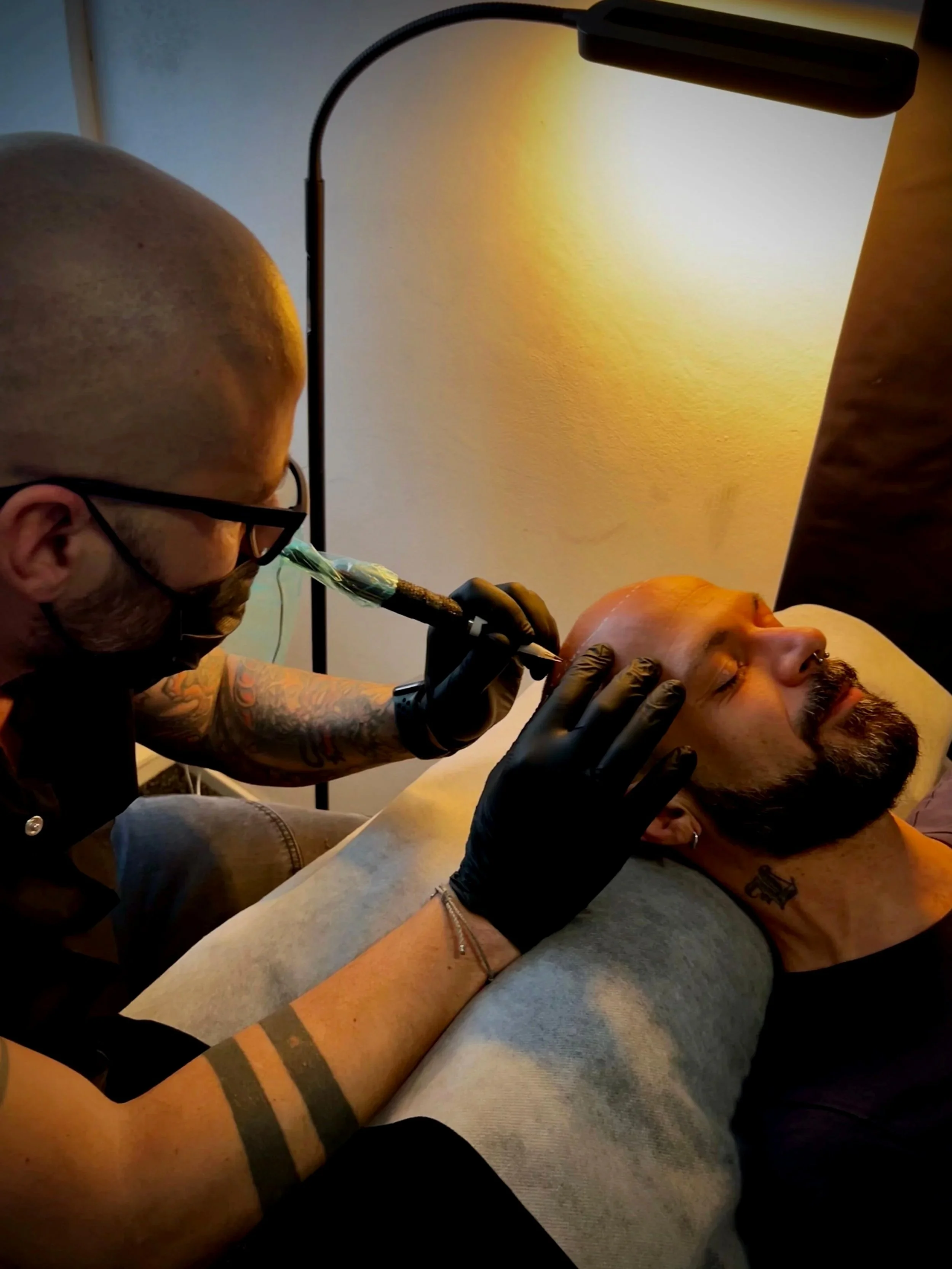 Un operatore estitico di tricopigmentazione esegue il trattamento di tatuoaggio capelli su un uomo sdraiato, con luci calde nell'ambiente.