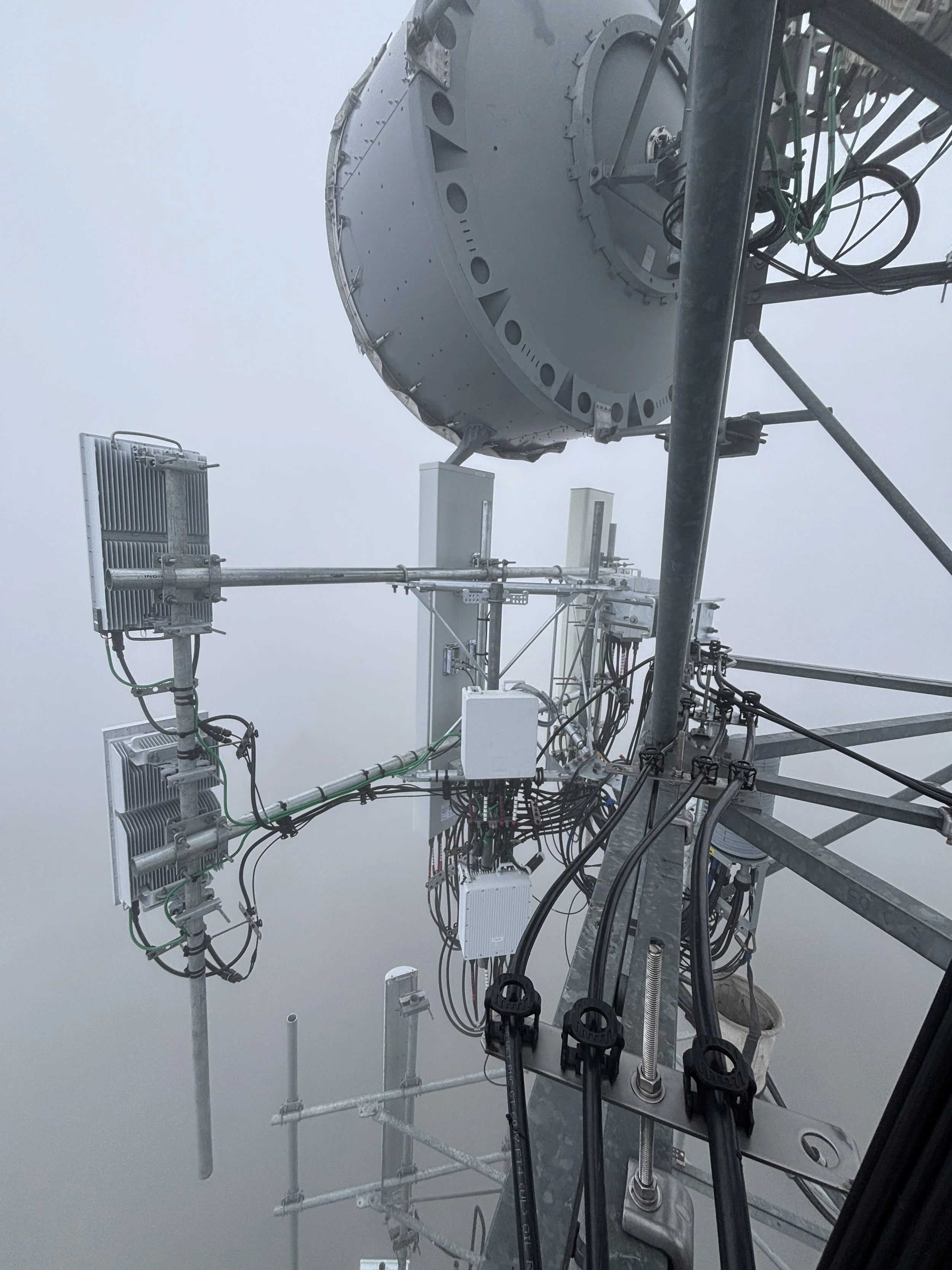 A close-up view of a telecommunications tower with various antennas and equipment, set against a foggy sky.