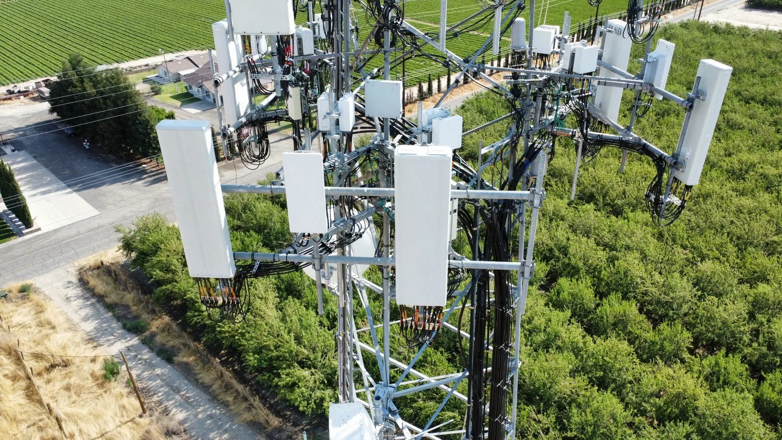 An aerial view of a large telecommunications tower with multiple antennas and equipment, surrounded by green foliage, roads, and nearby houses.