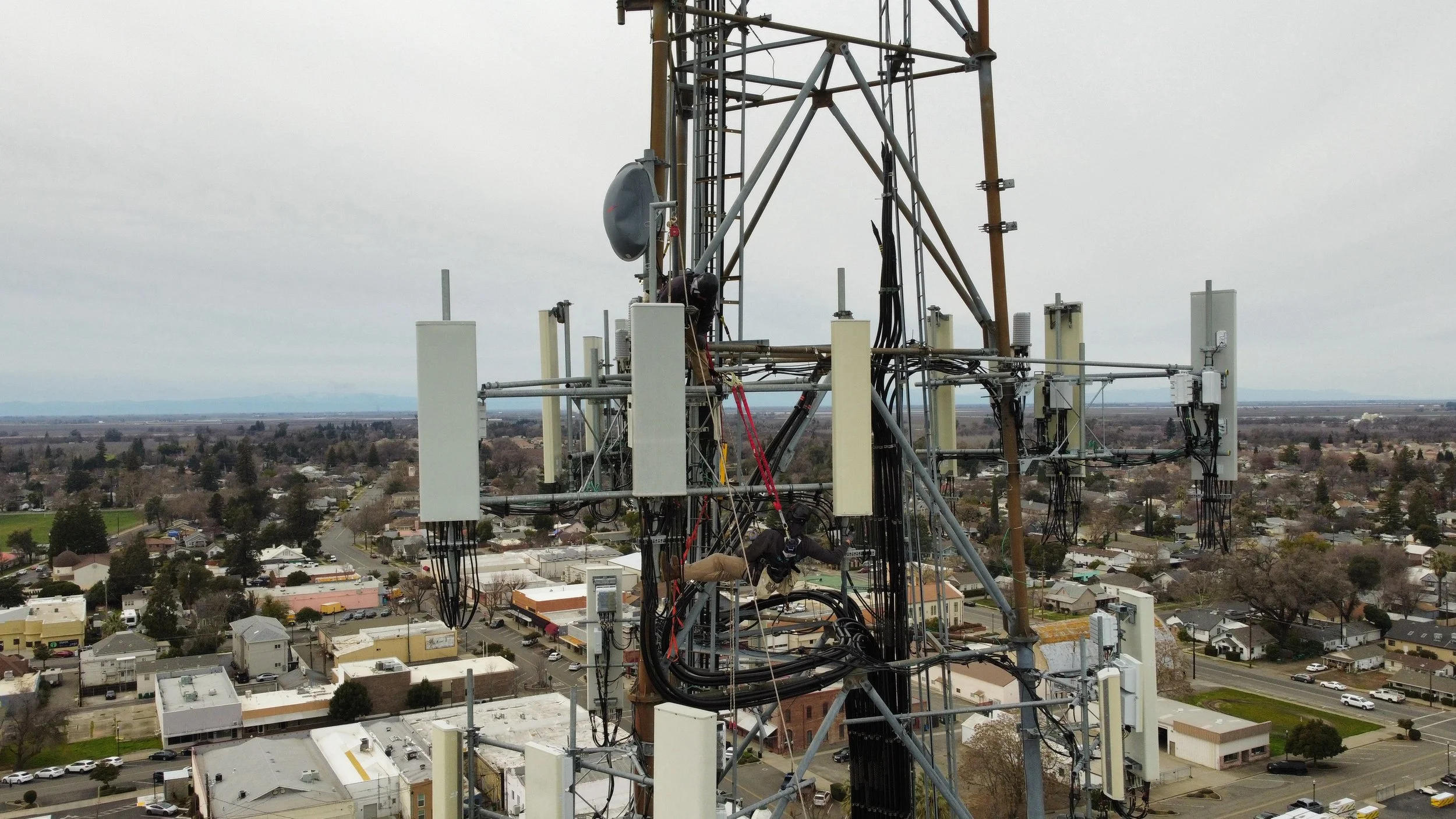 Technicians working on communication equipment on a tall radio tower above a suburban neighborhood during the daytime.