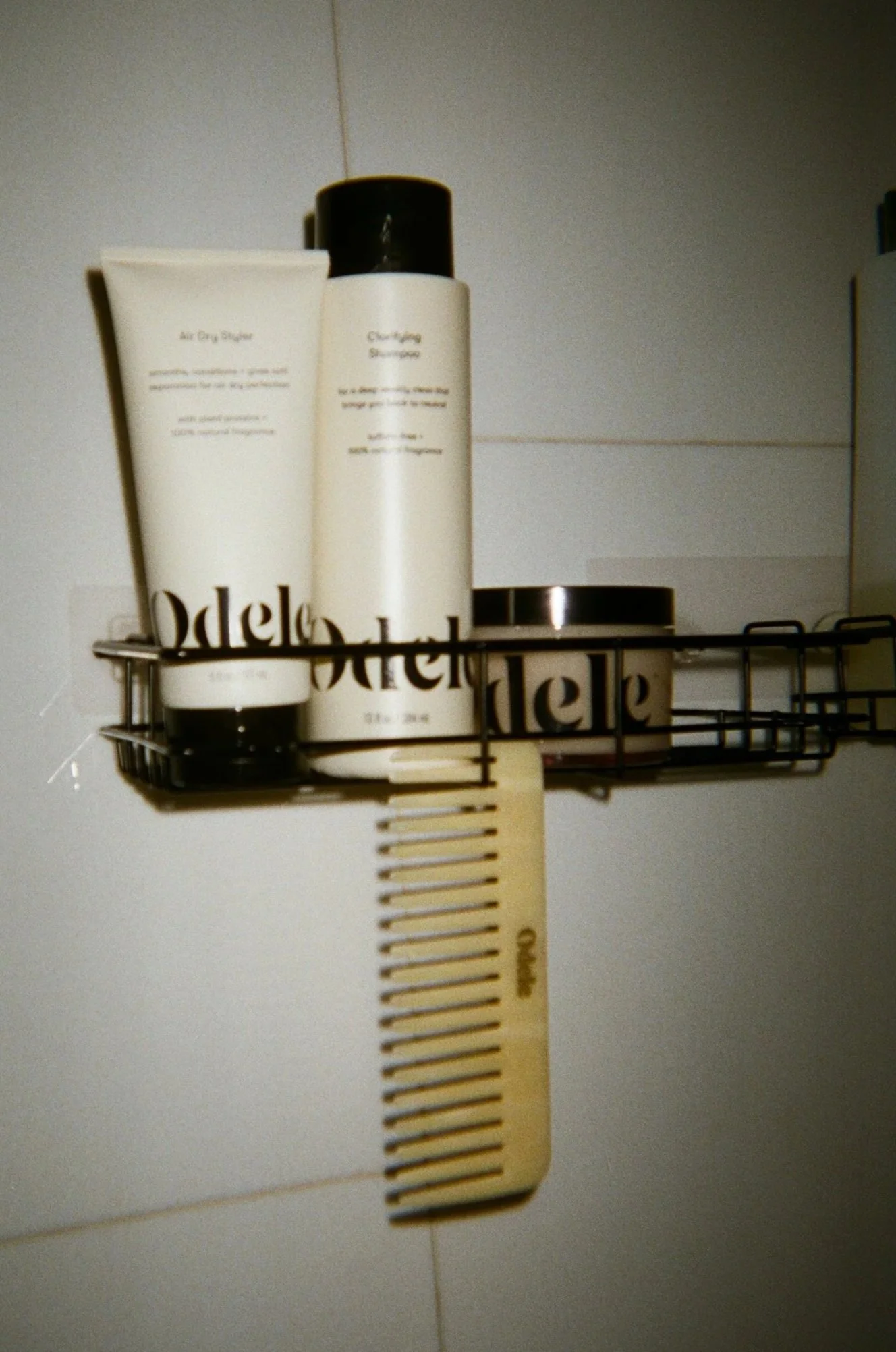 A metal shower caddy attached to a bathroom wall holding toiletries including two bottles of products, one labeled 'Air Dry Styler' and the other 'Curling Serum,' and a small jar.